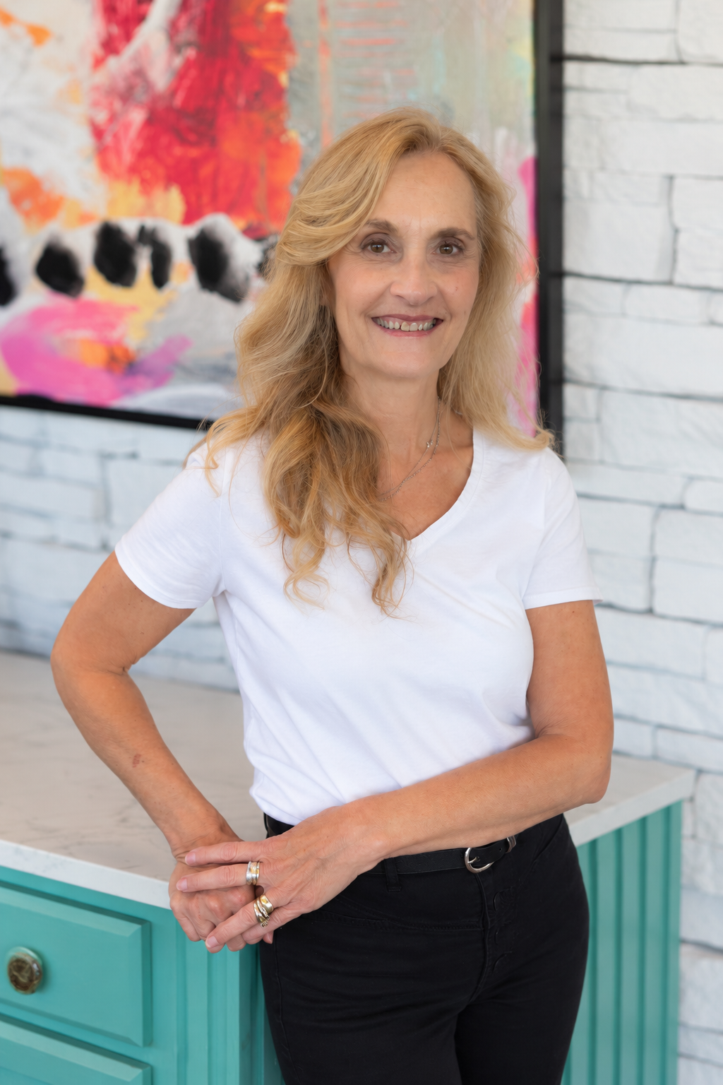 A woman with blonde hair smiling, wearing a white T-shirt and black pants standing indoors next to a turquoise cabinet with a white marble top, in front of a colorful abstract painting on a white brick wall.