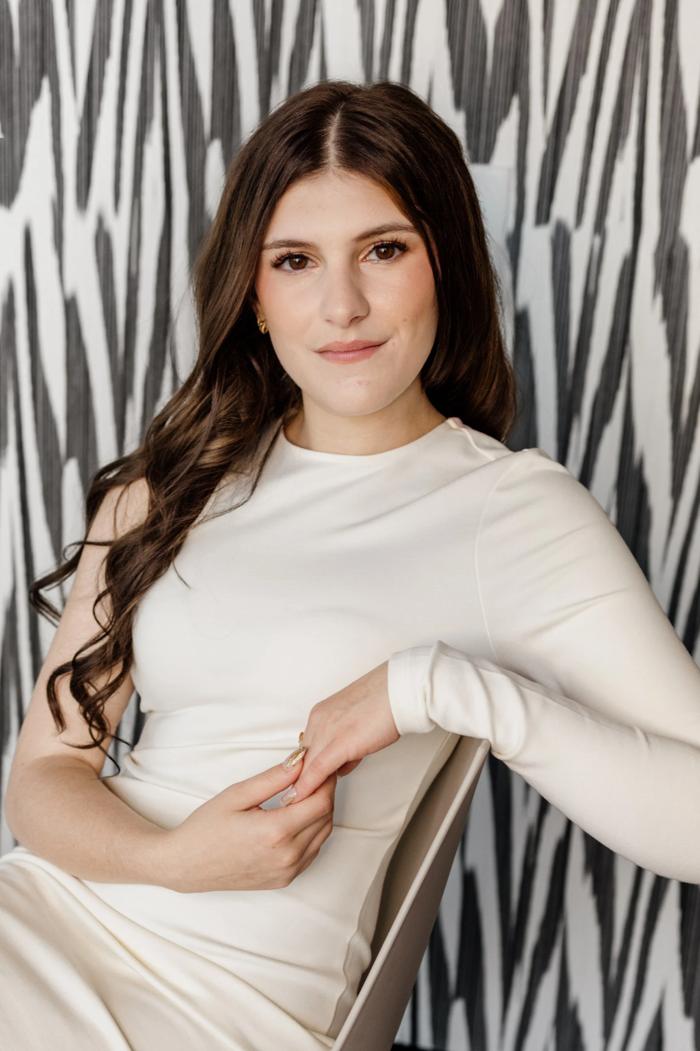 A woman with long, dark, wavy hair and light makeup, wearing a cream-colored long-sleeve top, sitting on a beige chair against a black and white abstract patterned background.