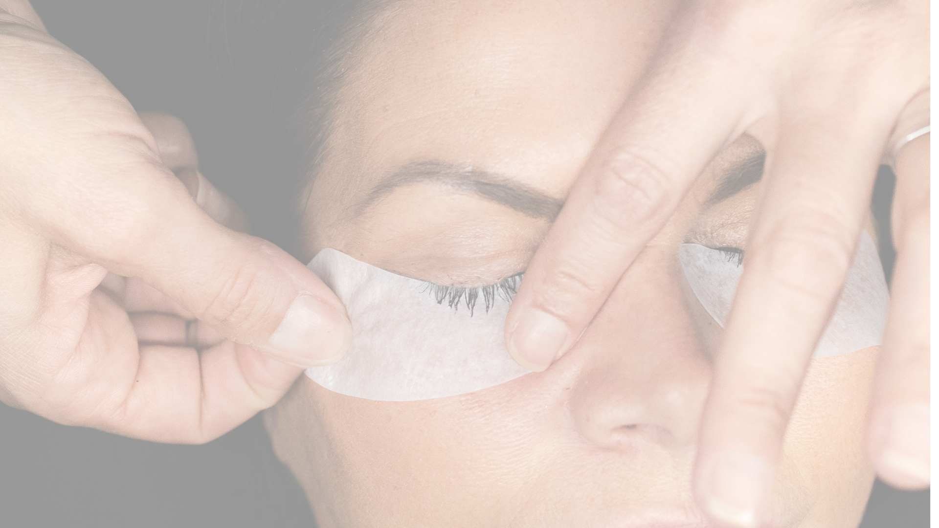 A woman receiving eyelash extensions from a technician in a salon or spa, with a close-up view of her face and the technician's hands applying the extensions.