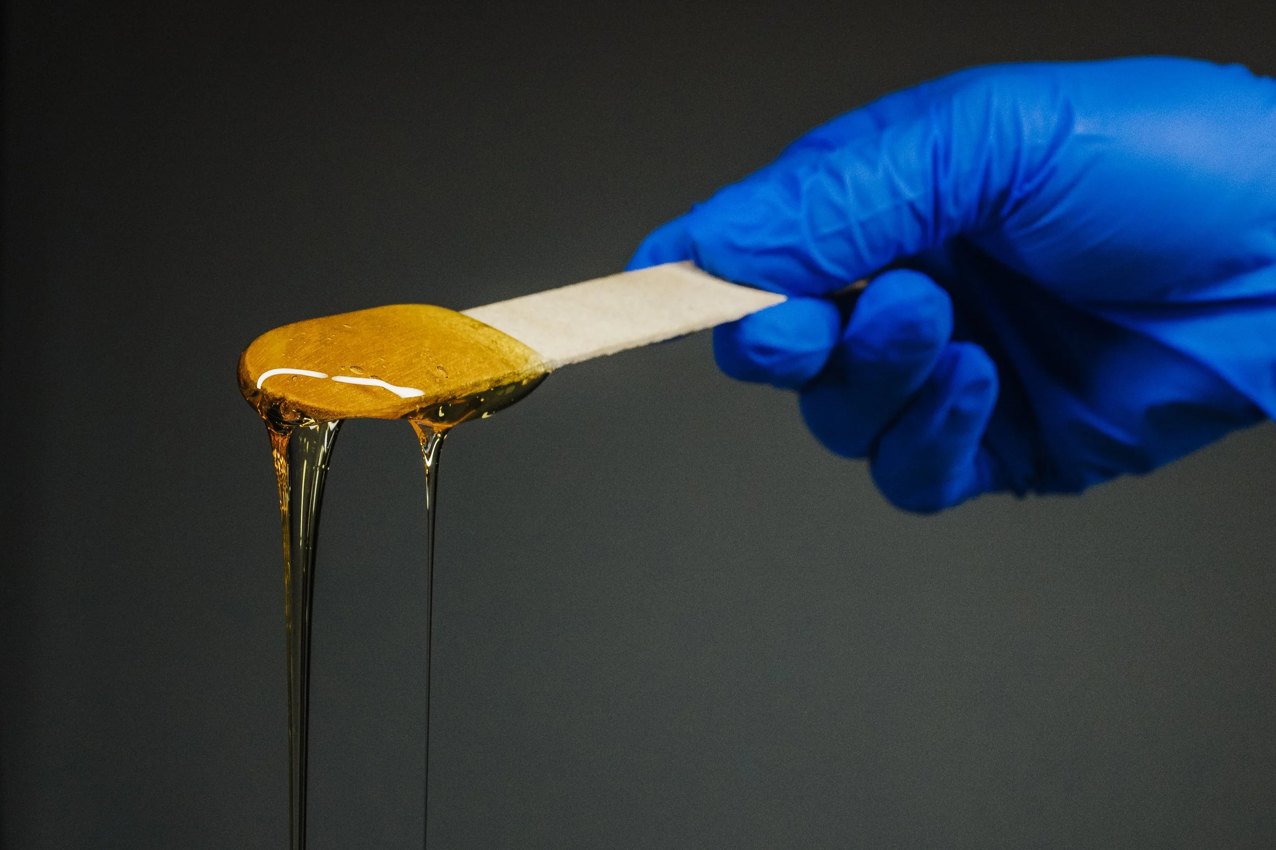 A gloved hand holding a wooden spatula with honey dripping from it.