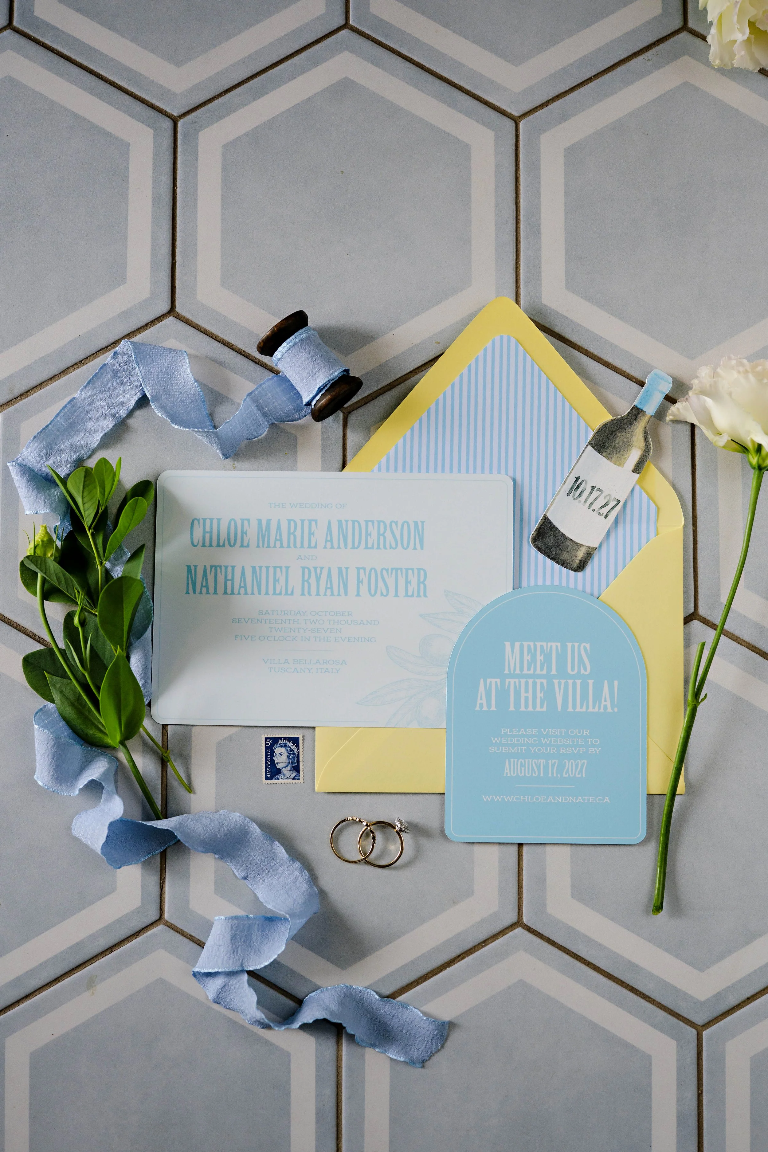 Wedding invitation, flowers, and rings on a tiled floor, with a blue ribbon, postage stamp, and a bottle-shaped tag with the date 10.17.27.
