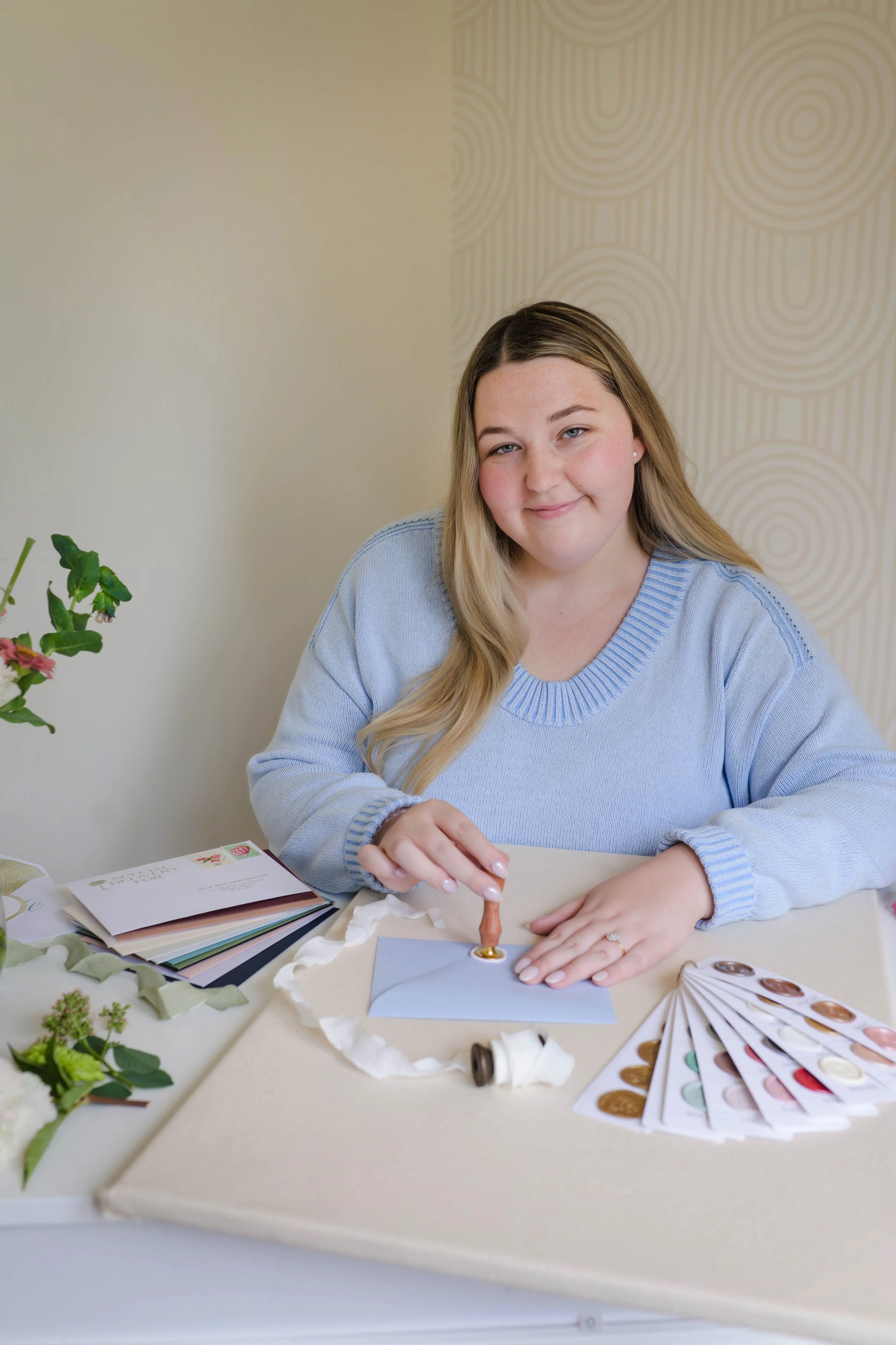 A woman with long blonde hair wearing a light blue sweater, sitting at a table with various color swatches, paint samples, and decorative items, holding a small object over a piece of paper, with a beige patterned wall and a small green plant to her left.