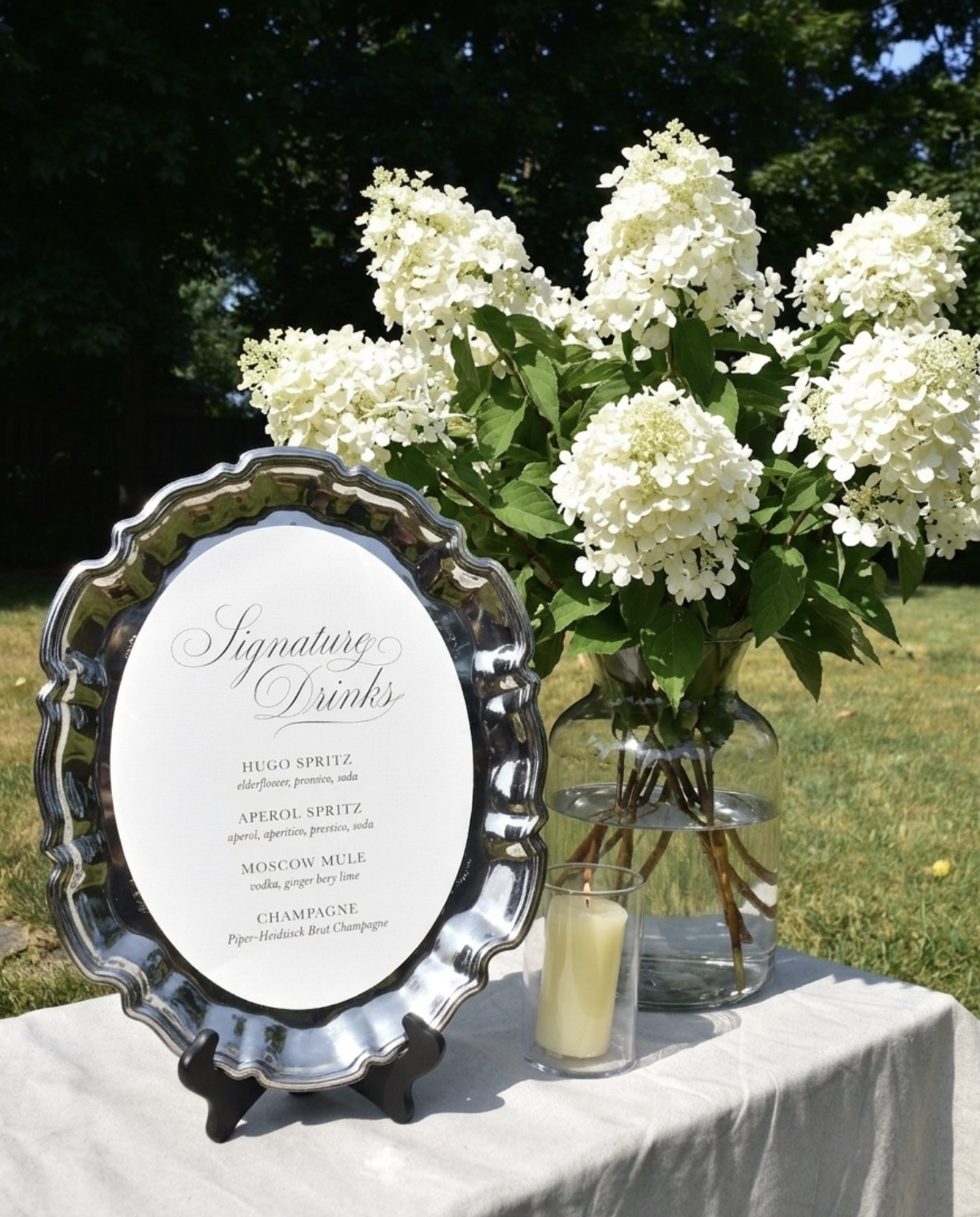 A table setting outdoors with a silver-framed menu listing signature drinks, a large glass vase with white hydrangea flowers, a small candle in a glass holder, and a white tablecloth, with trees and blue sky in the background.