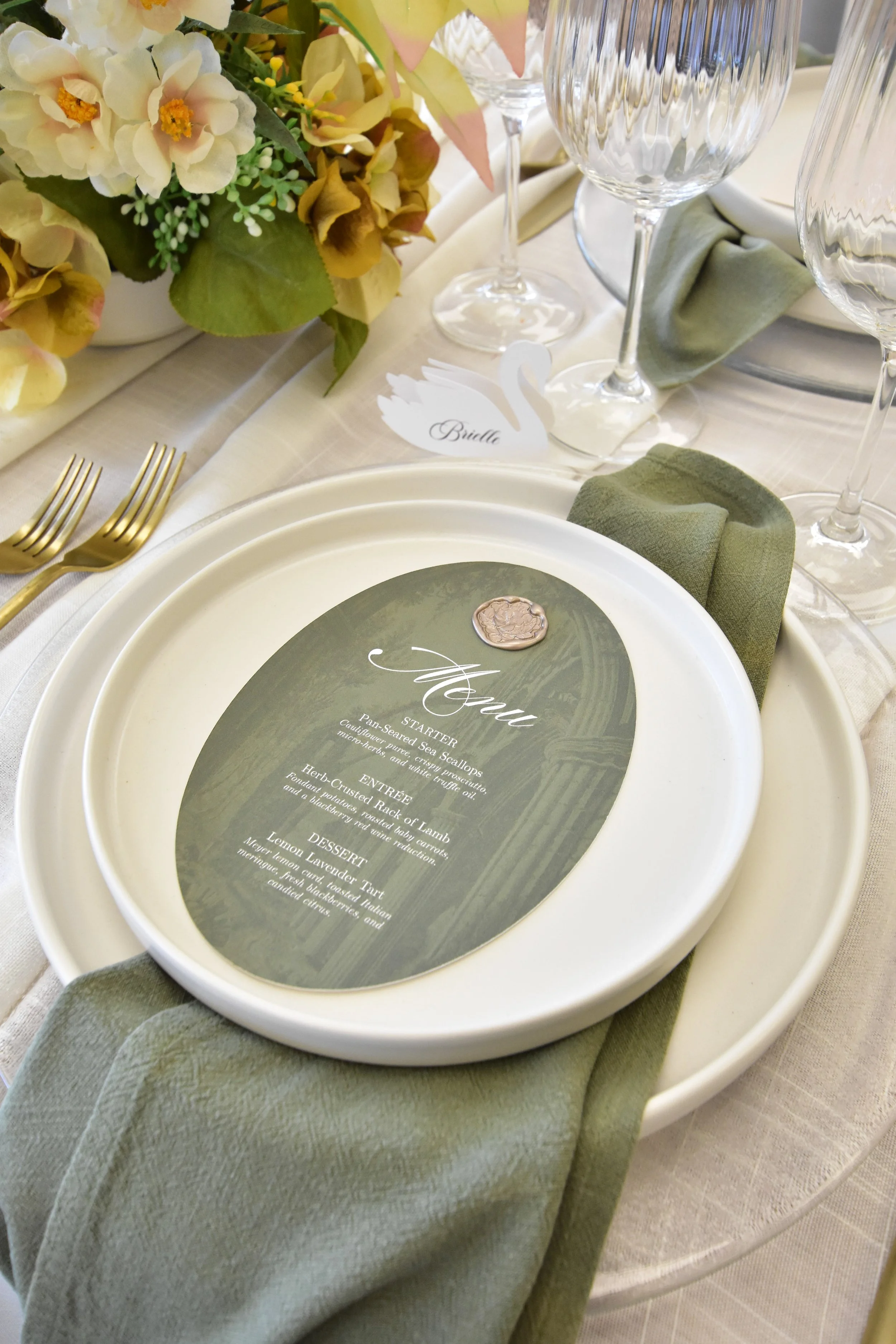 A formal table setting with a floral centerpiece, empty wine glasses, and a menu placed on a plate. A green cloth napkin is folded beside the plate. There is a small decorative swan name card with the name 'Brielle' on it.
