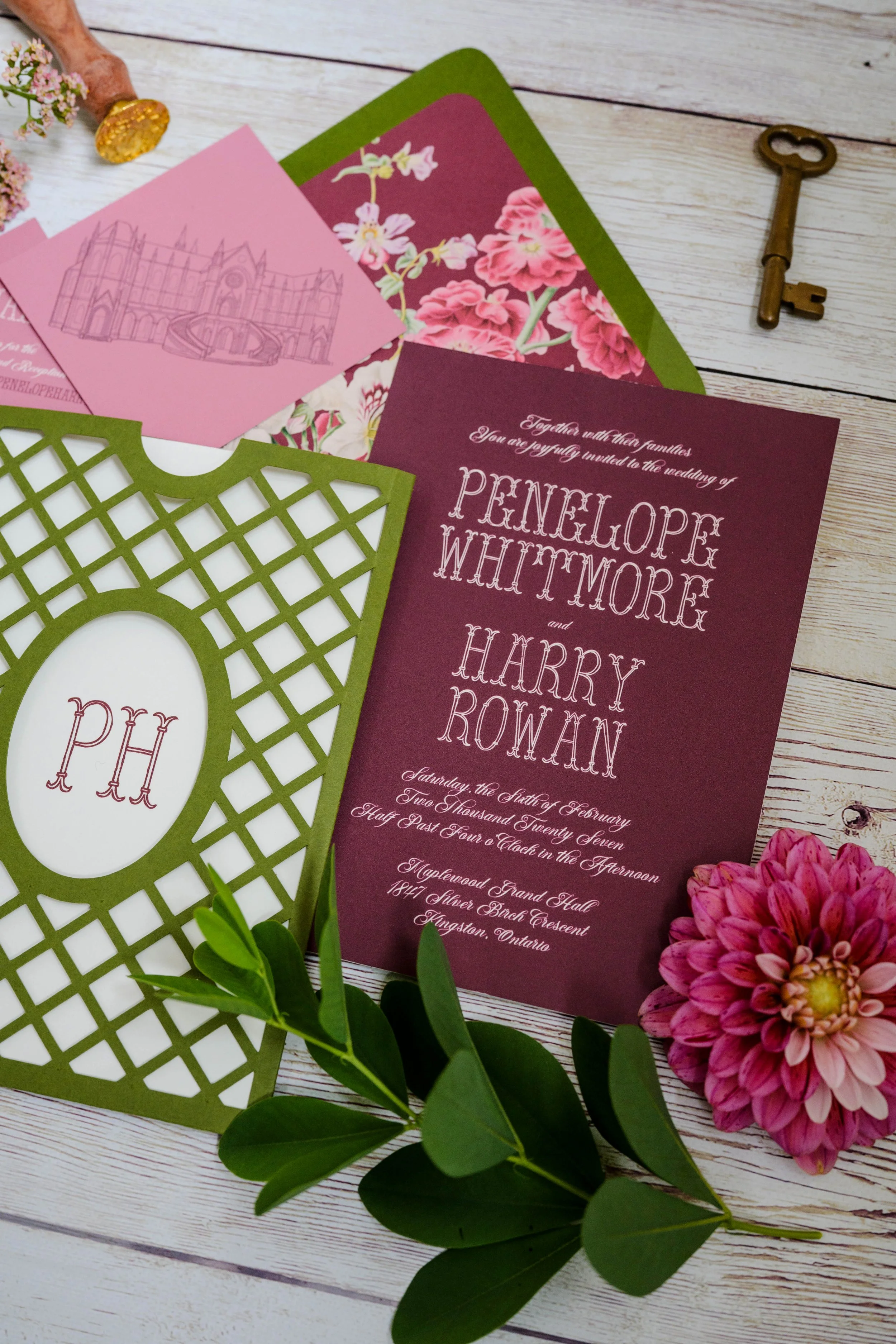 Wedding invitations with floral designs, a green lattice photo frame, a large pink flower, a gold-colored key, and decorative pink and white flowers on a white wooden surface.