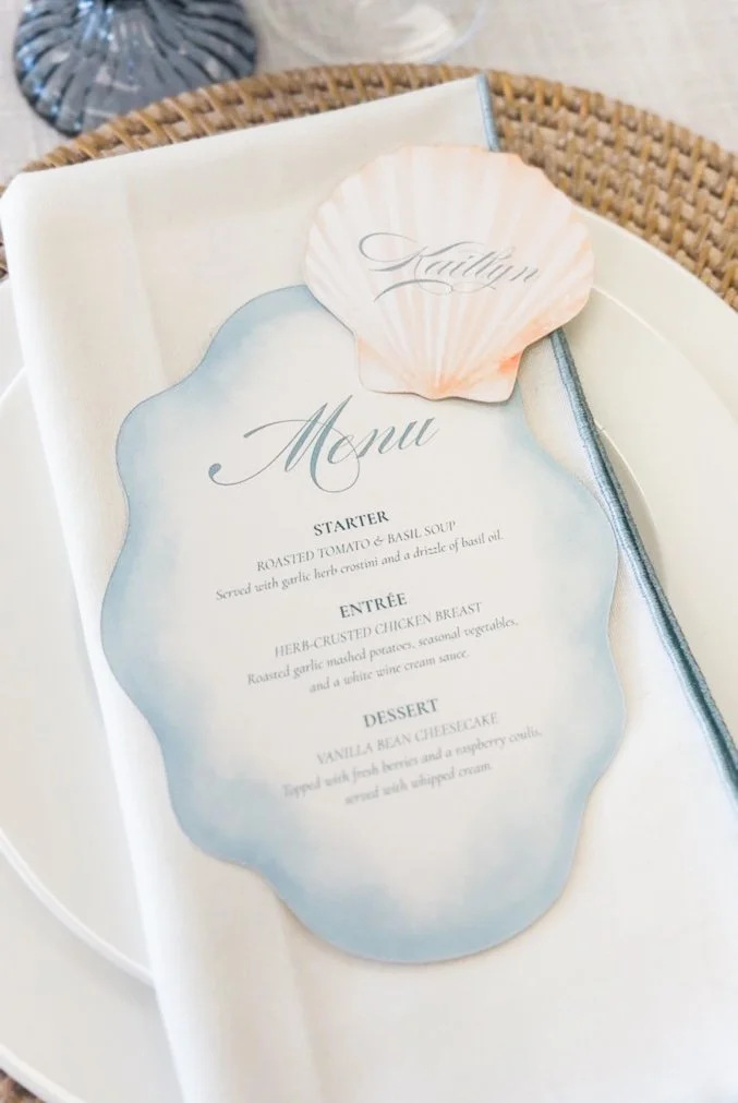The image shows a wedding menu placed on a white table, with a pink scallop shell on top. The menu is decorated with a watercolor-style background and contains three sections: starter, entrée, and dessert, with descriptions of each dish. The sections are labeled 'Kaitlyn' and 'Menu' with elegant script fonts.
