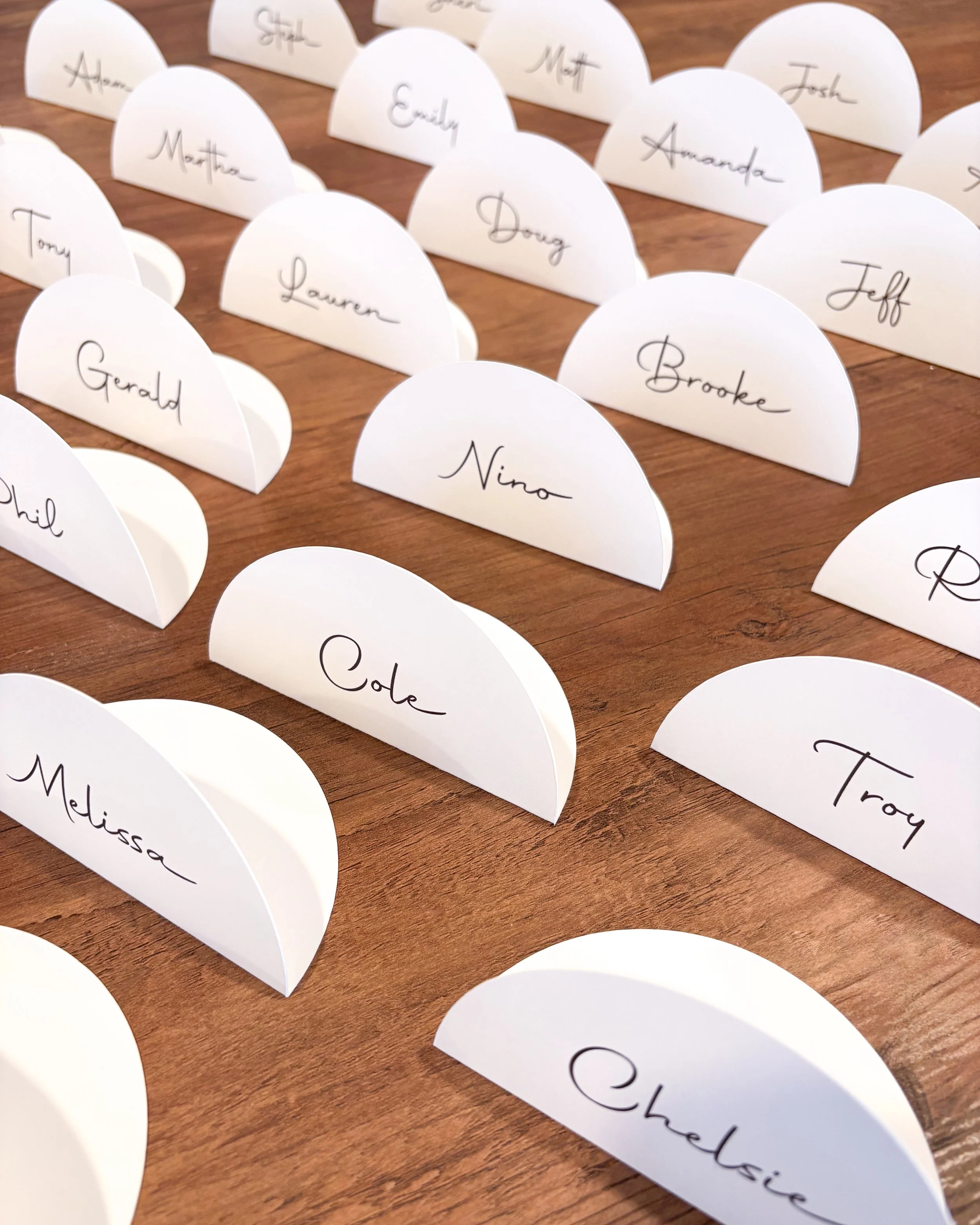 White semicircular place cards with handwritten names placed on a wooden surface, part of a seating arrangement for an event.