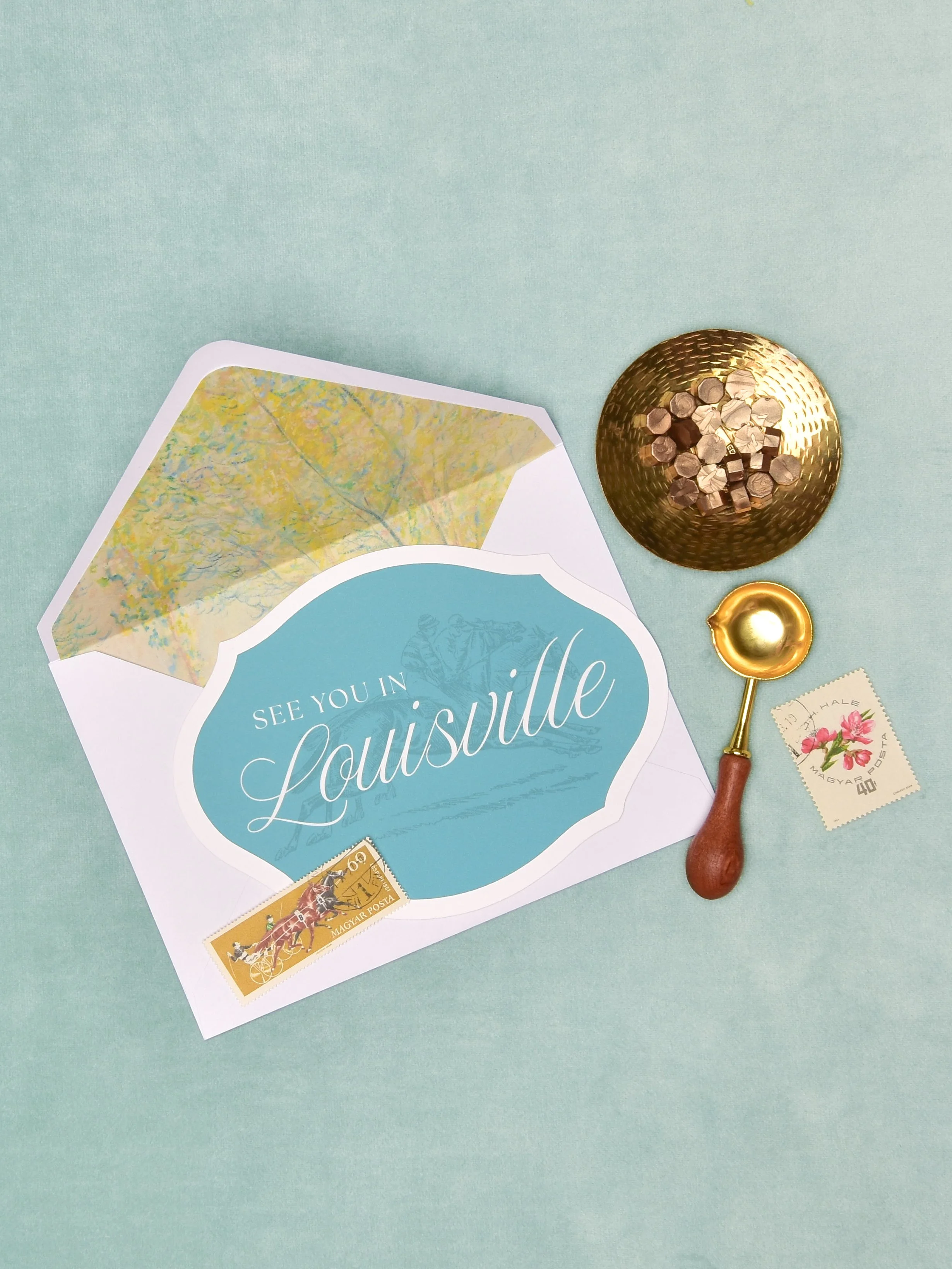 A Save The Datewith the text "See you in Louisville," a small pile of wax seal chips, a gold spoon, a postage stamp with pink flowers, and a small yellow postage stamp with a horse and carriage, all on a pastel blue background.