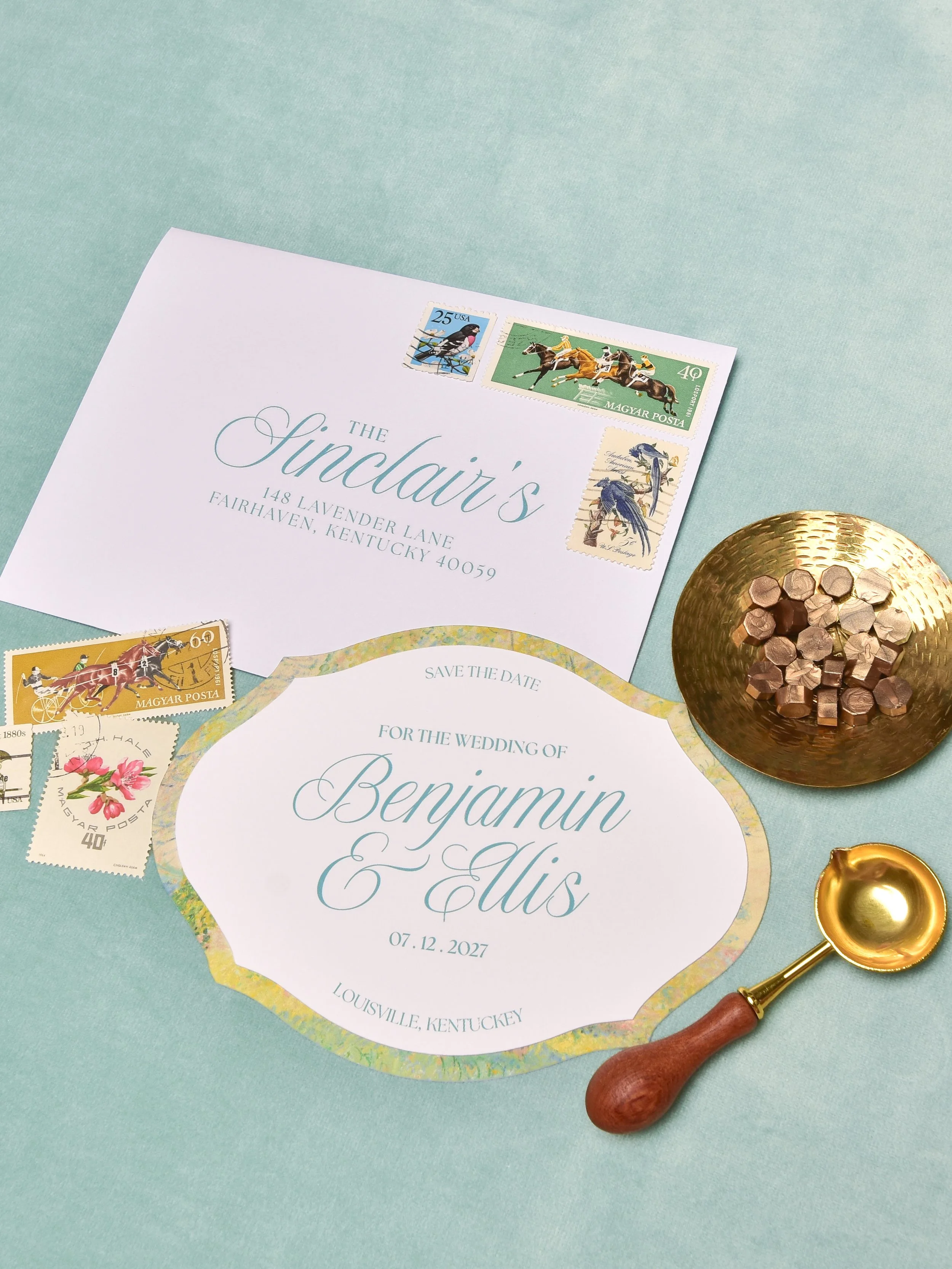 Wedding invitation with address and date details, surrounded by vintage postage stamps, gold bowl filled with wax seal chips, and a gold spoon with a wooden handle on a light blue background.
