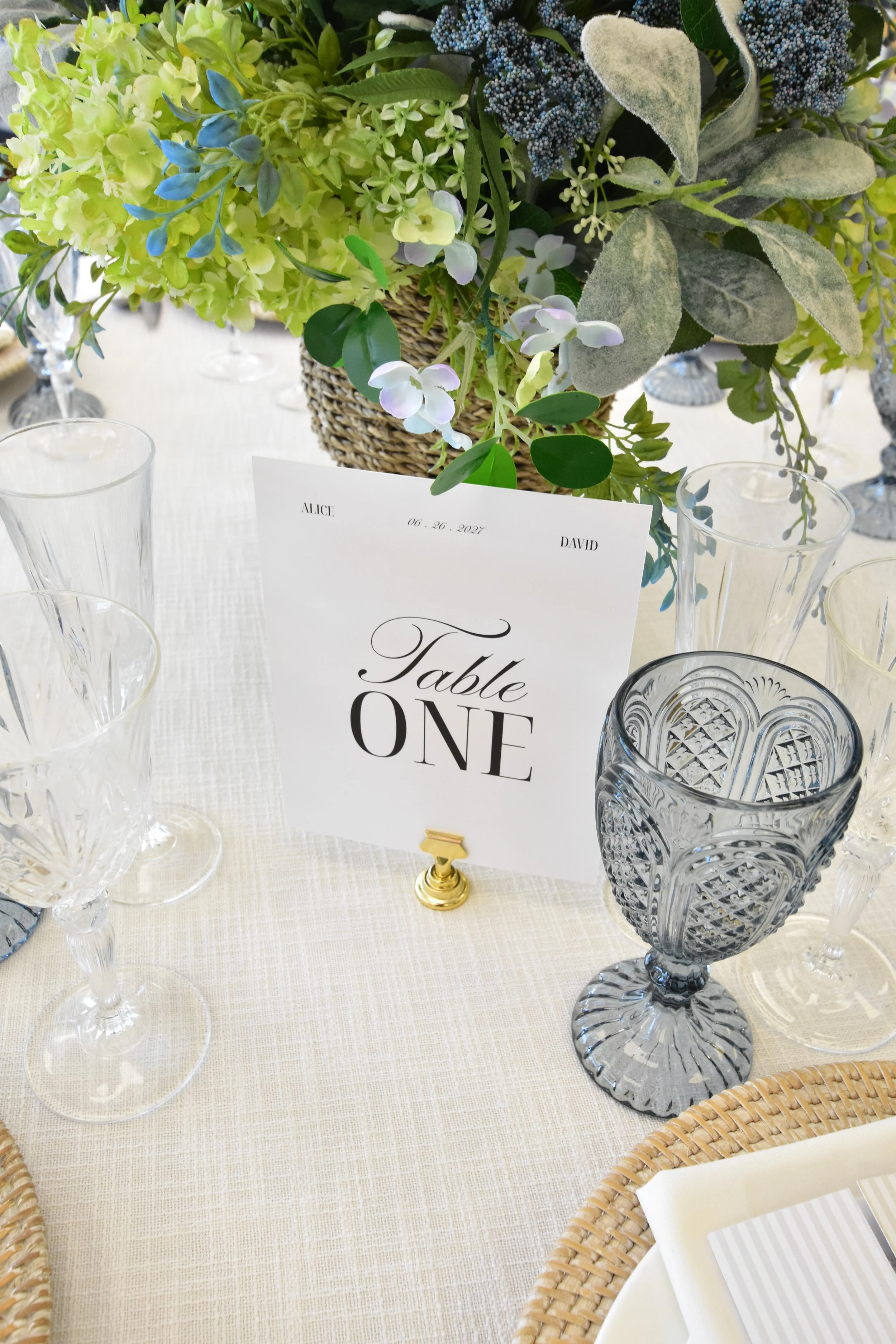 A tablescape decorated with an arrangement of green, white, and purple flowers in a wicker basket, surrounded by empty crystal glasses and a table sign with the date June 26, 2027, for a formal event.