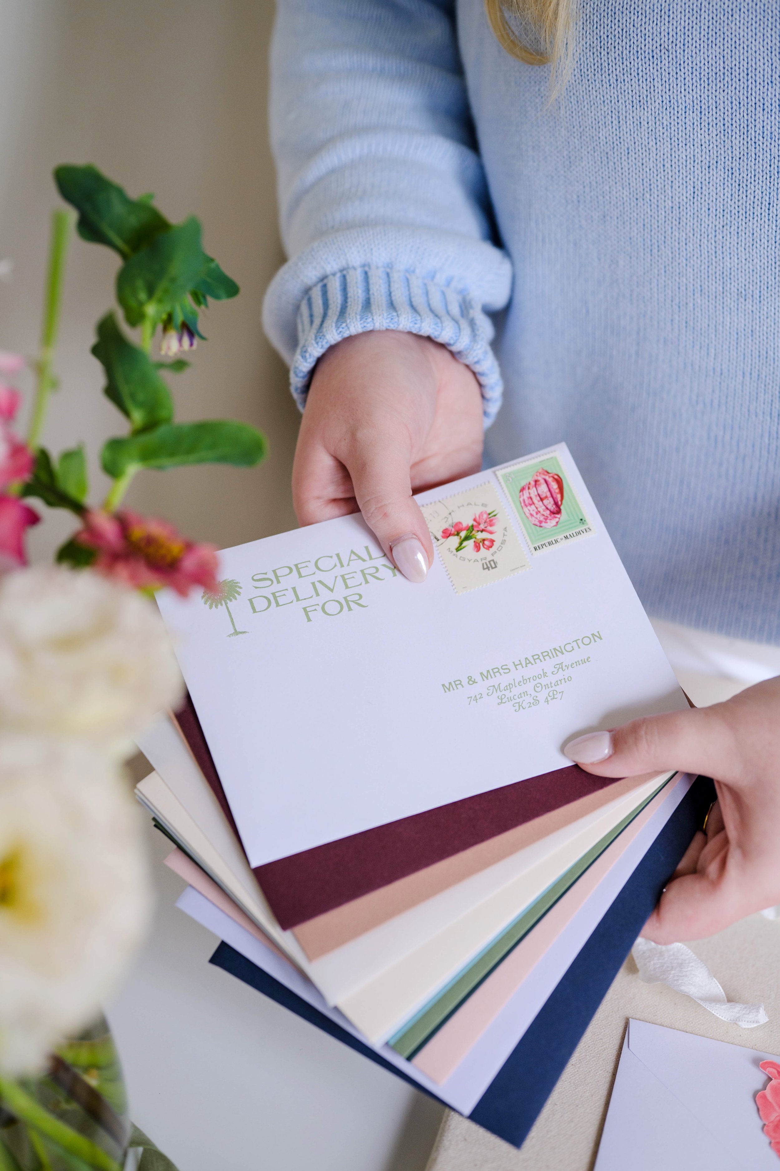 Person holding an envelope with printed addresses and postage stamps, standing next to a bouquet of flowers.