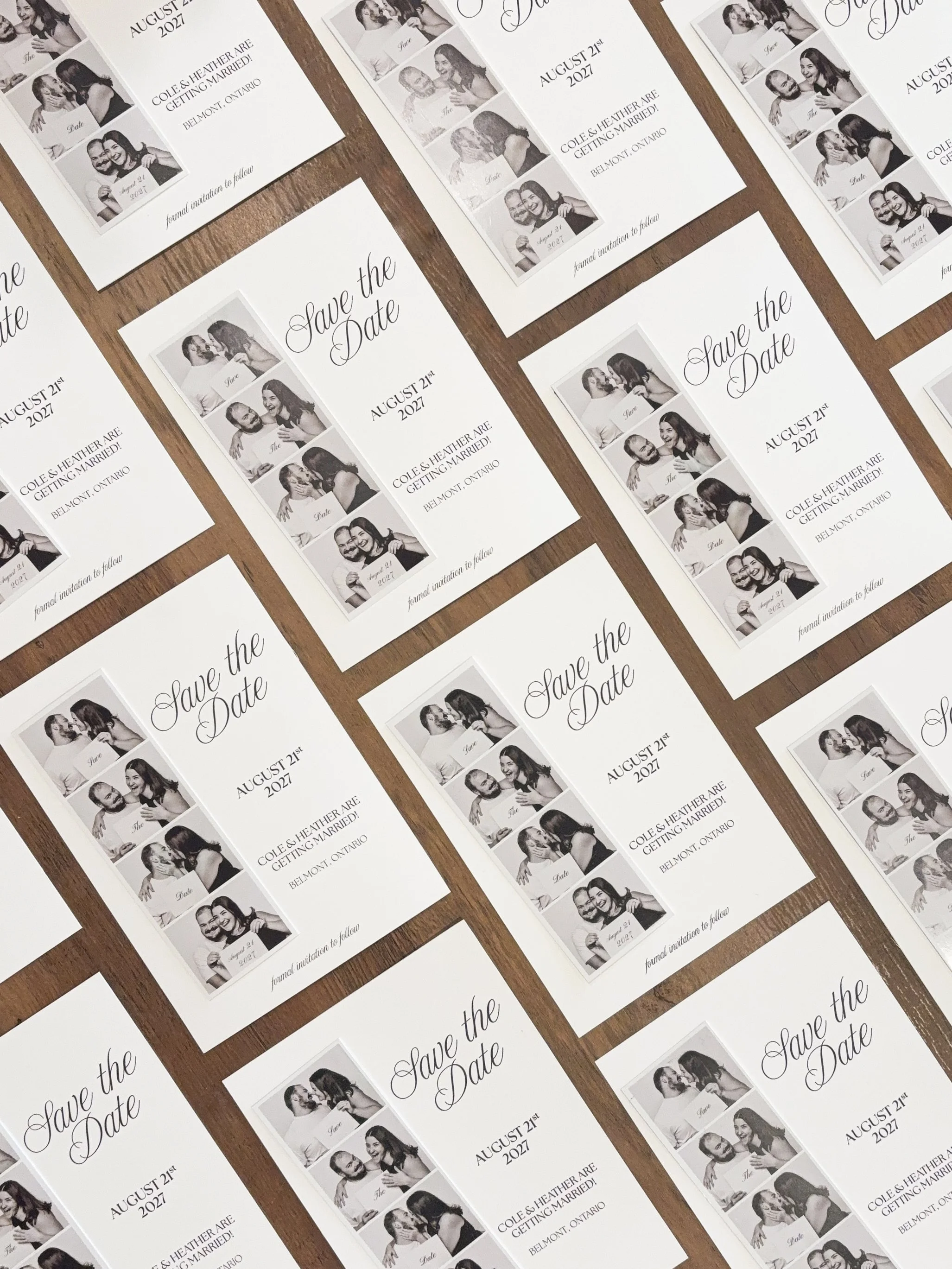 Multiple printed wedding save-the-date cards with black-and-white photo strip and text indicating the event date as August 21, 2027, and location in Belmont, Ontario, arranged on a wooden surface.