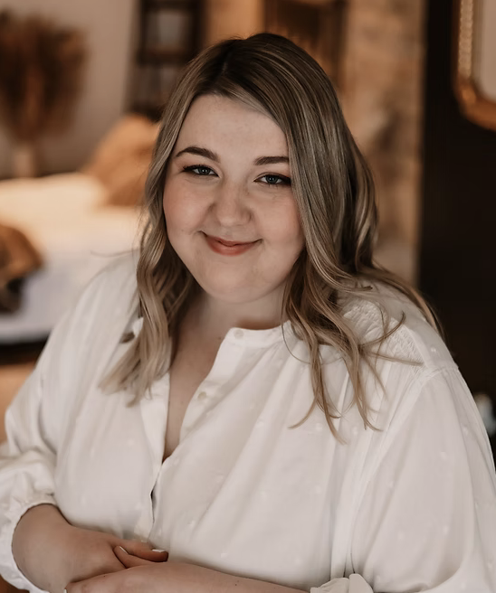 A young woman with shoulder-length wavy blonde hair, wearing a white blouse, smiling in a cozy indoor setting with brick walls and warm lighting.