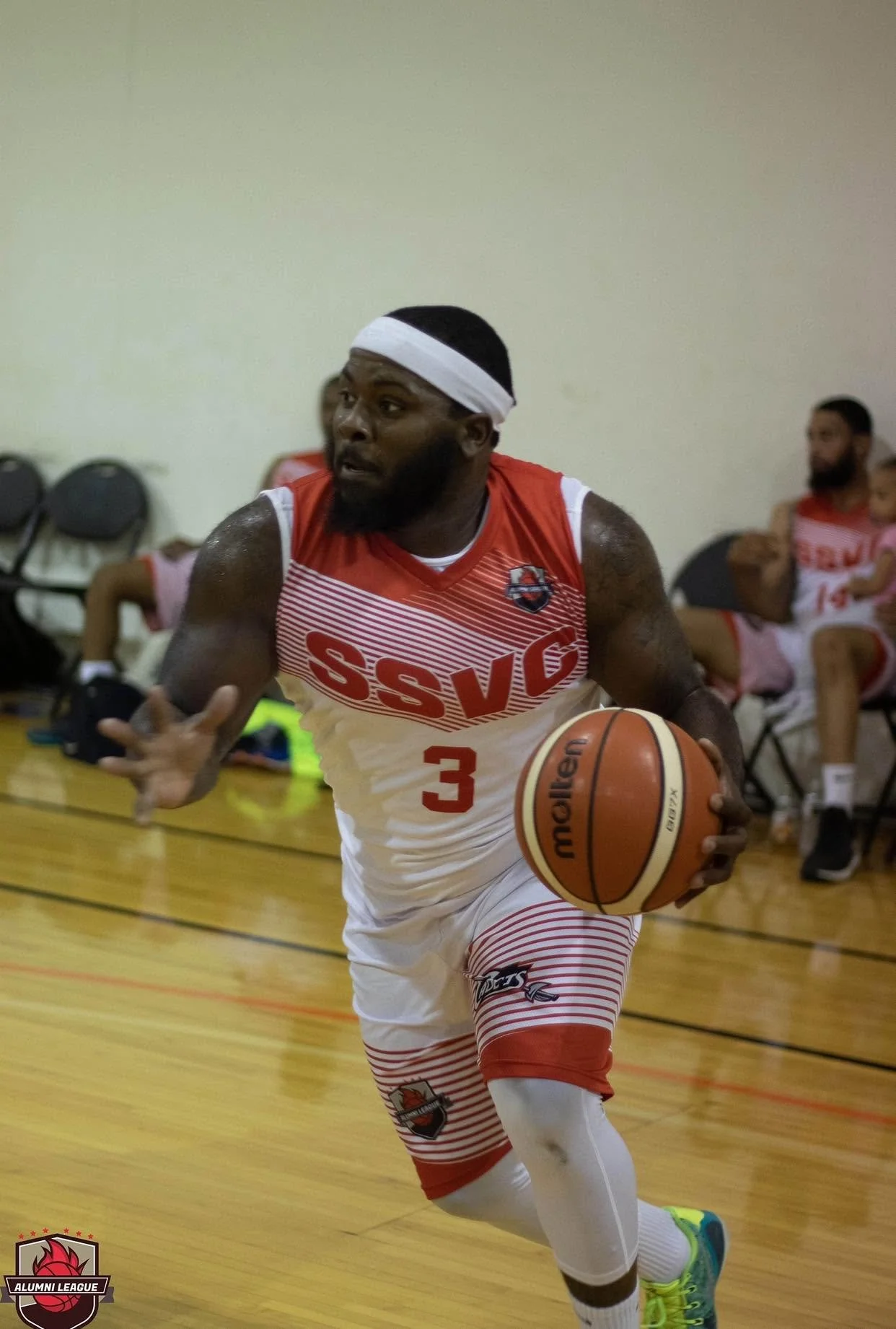 A basketball player wearing a red and white jersey with the number 3 dribbling a basketball on an indoor court. The player has a white headband and is running with a focused expression. In the background, there are other players sitting on chairs.