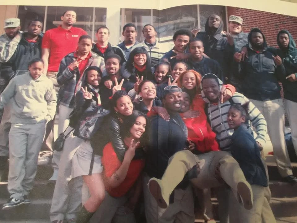 A group of young people and adults posing together in front of a building, some making peace signs or gestures, smiling and joyful.