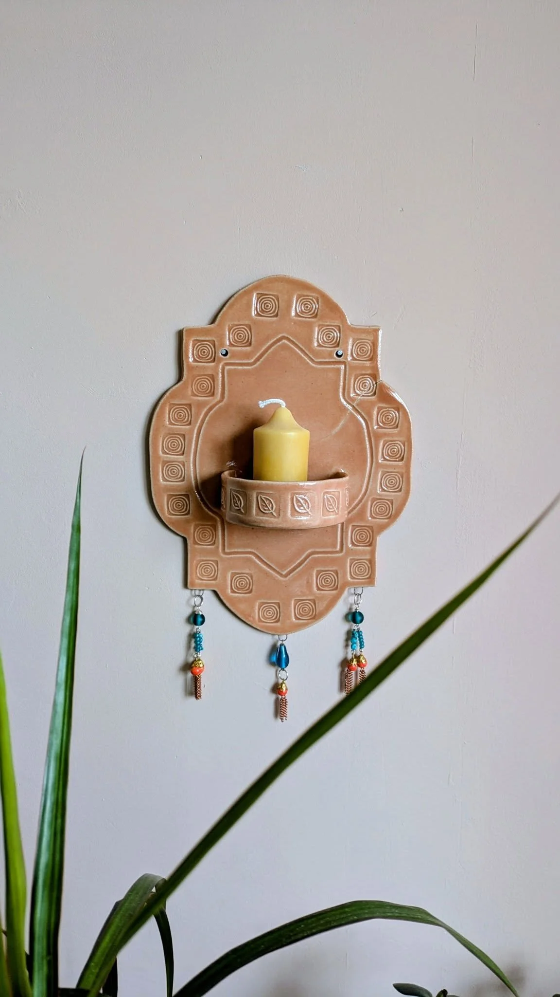 Handbuilt terracotta wall-mounted candle holder with geometric patterns and colorful hanging beads, holding a yellow pillar candle against a neutral wall.