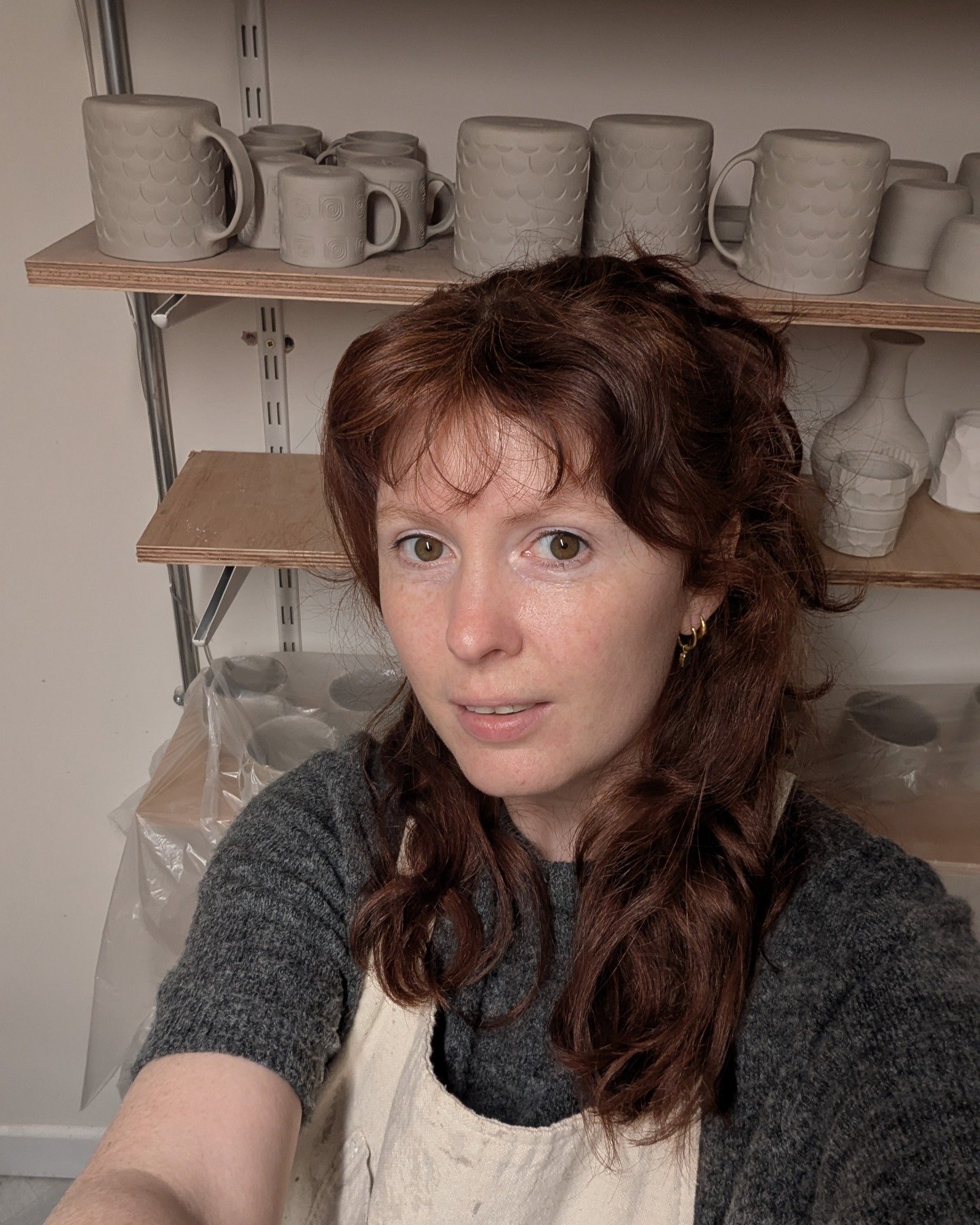 Kaira in the Fireweed Pottery studio wearing an apron, with handmade mugs and vases on shelves behind her.