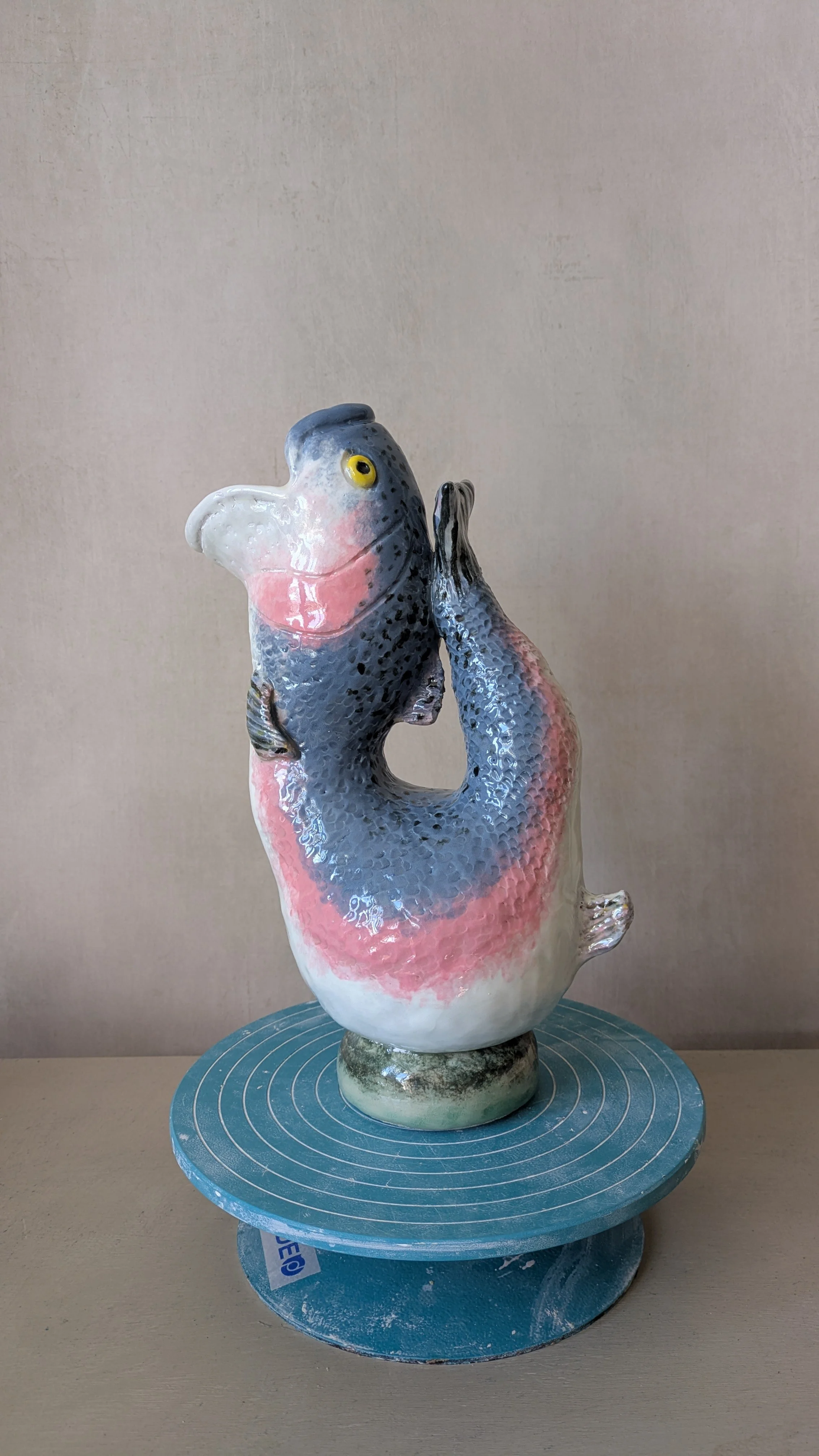 Handbuilt ceramic gluggle jug sculpture shaped like a fish, finished with blue, pink, and white glaze, displayed on a pottery wheel.