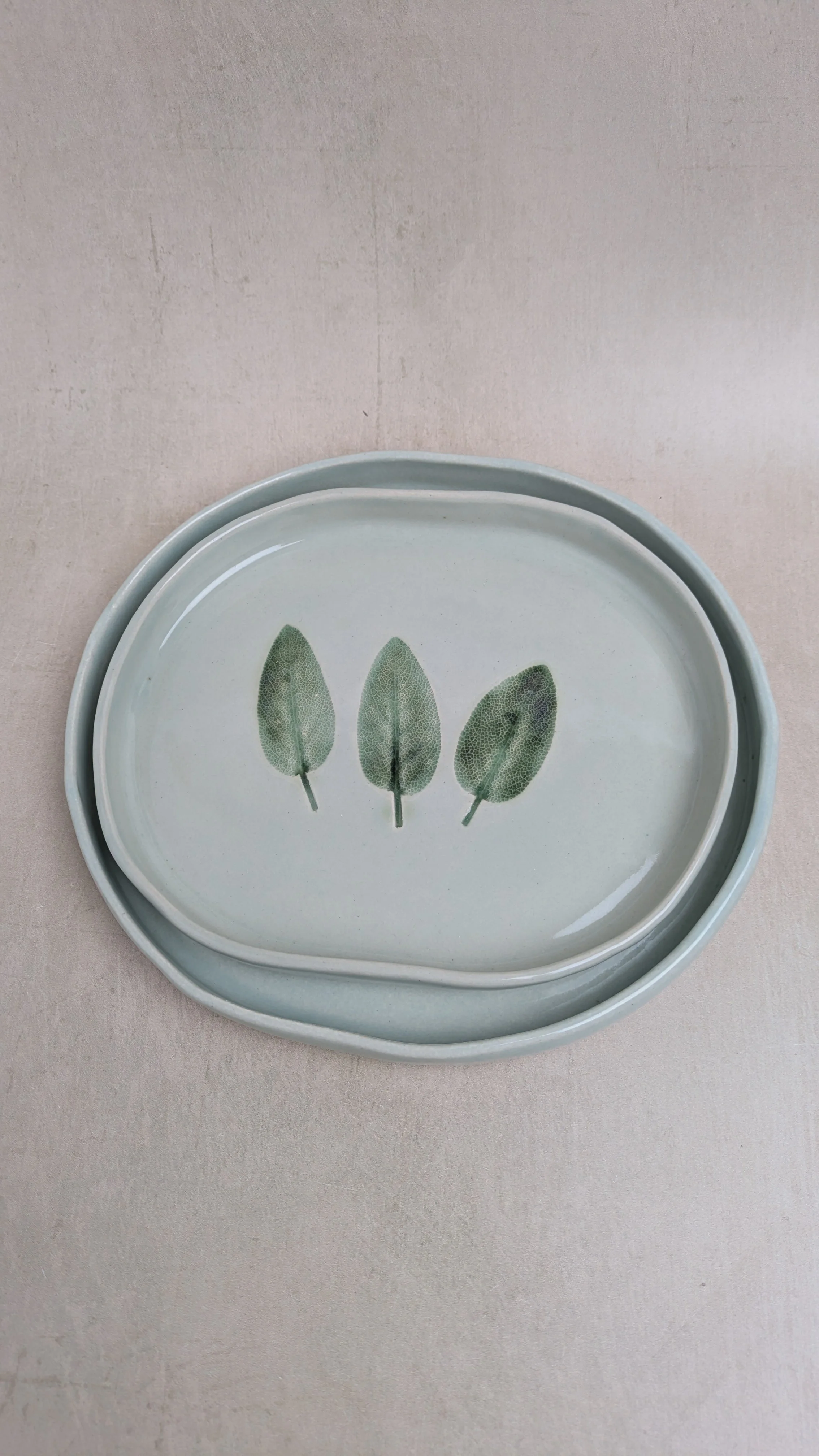 Stacked oval stoneware plates in sage glaze with sage leaf imprints. Placed on olive wood display