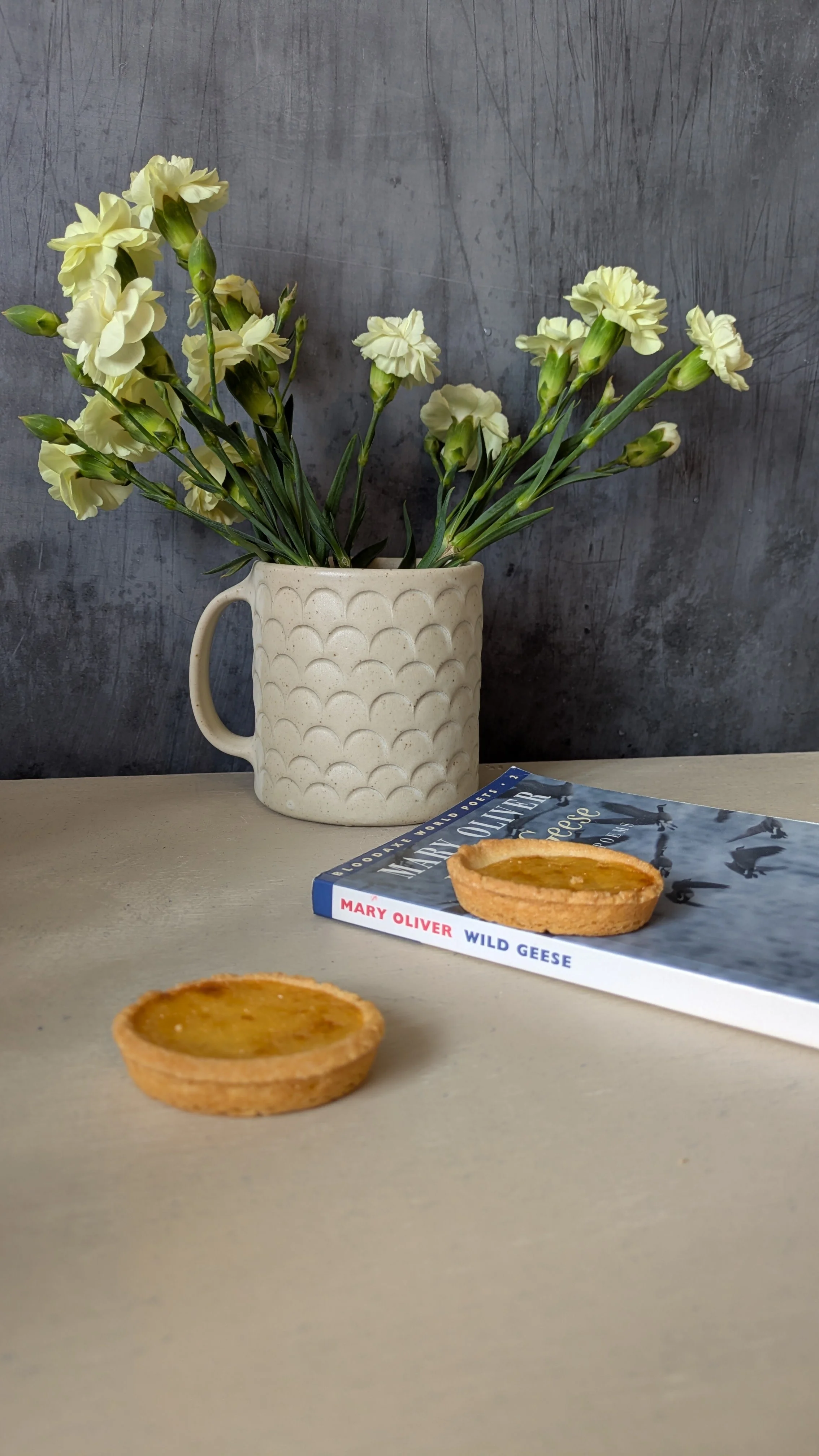 Large cream textured mug handmade at Fireweed Pottery, used as a vase with pale yellow carnations beside a book and tart on a neutral surface.