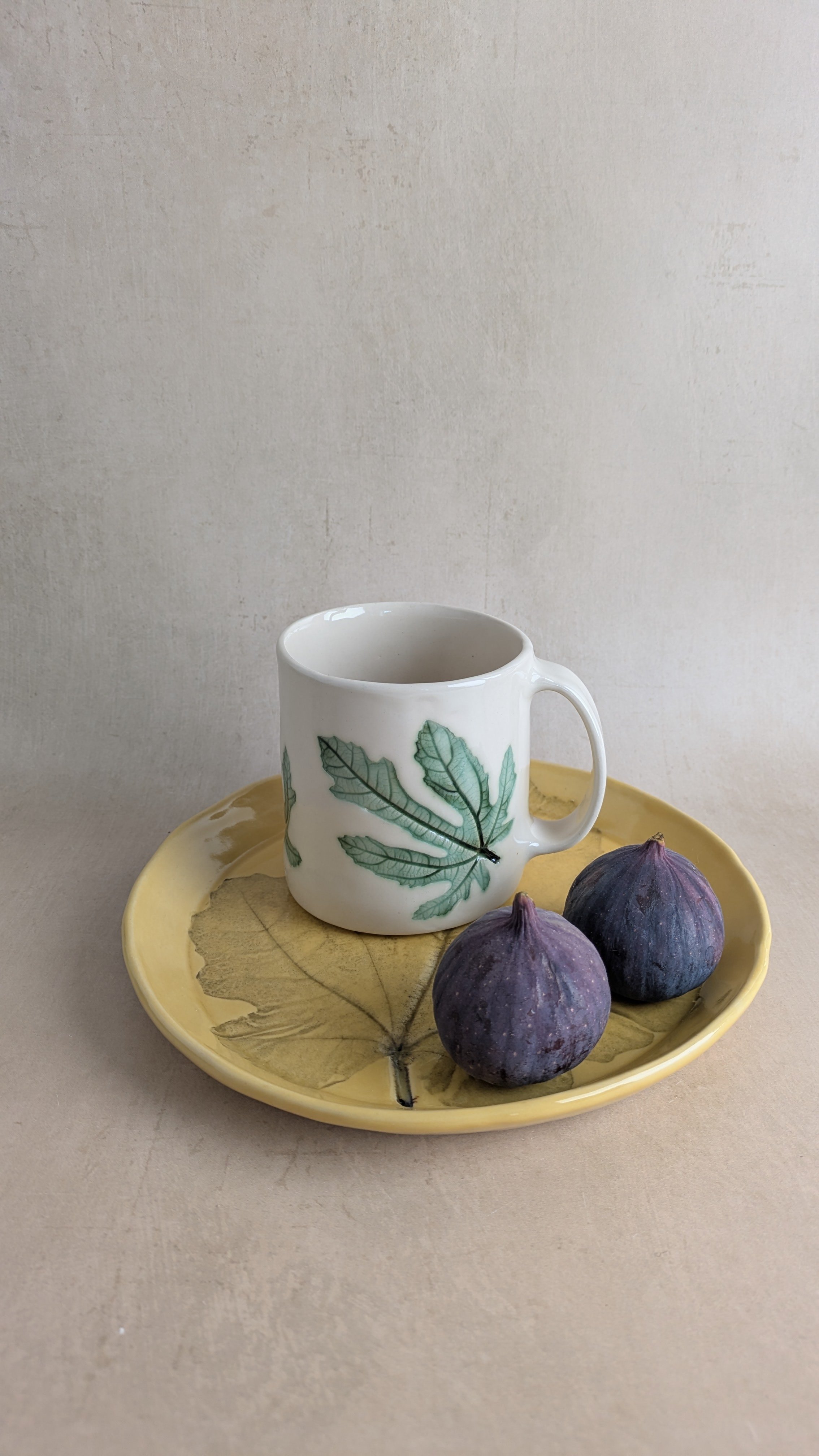 Handmade yellow‑glazed stoneware plate with fig‑leaf imprint, paired with a white mug and two fresh figs.