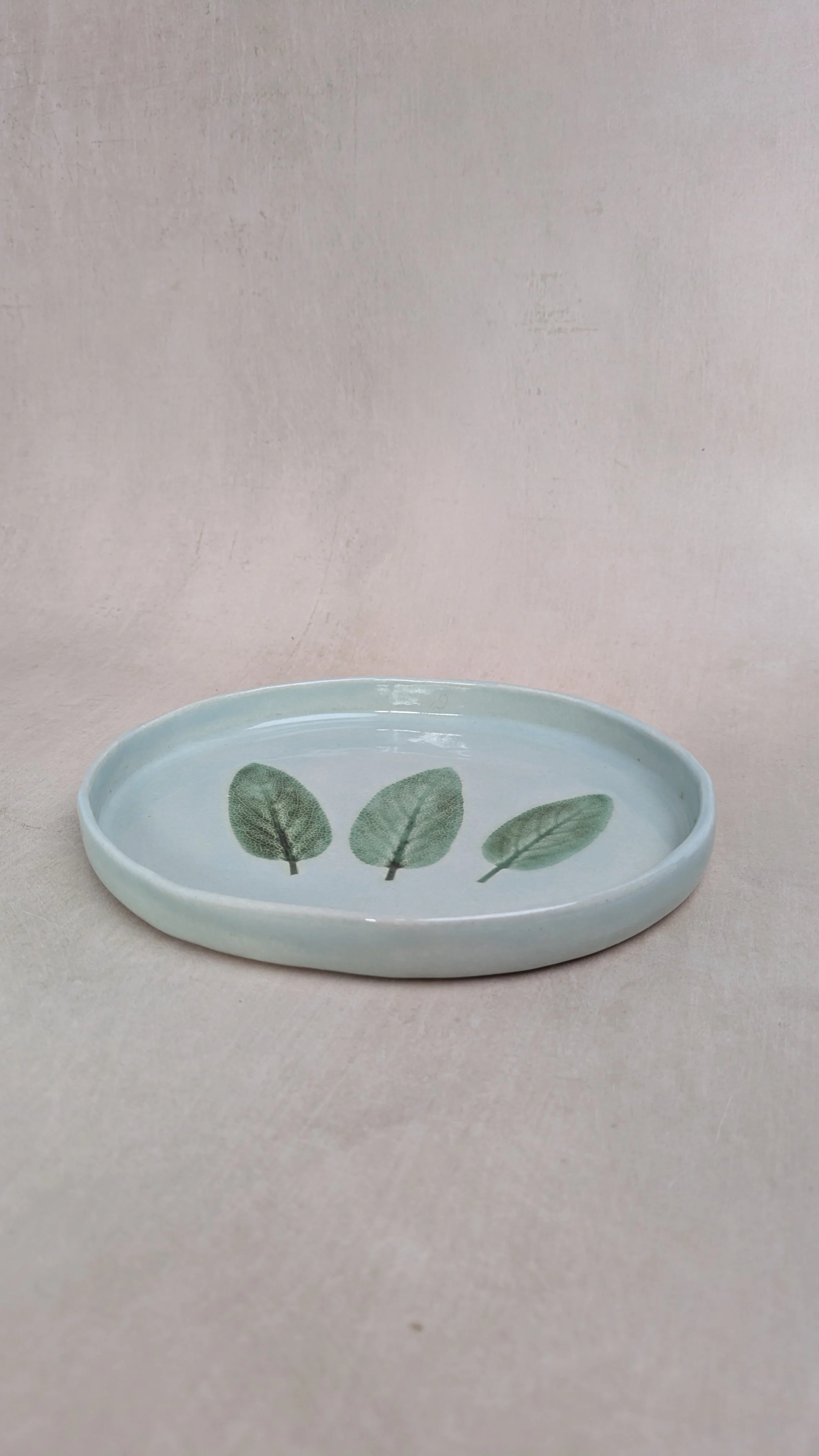 Oval stoneware plate with three sage leaves pressed into pale blue glaze, shown from side angle