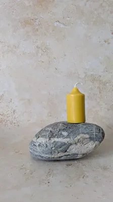 Hand‑poured yellow beeswax pillar candle resting on a smooth grey stone with white streaks, set against a soft beige textured background for a simple, natural composition.