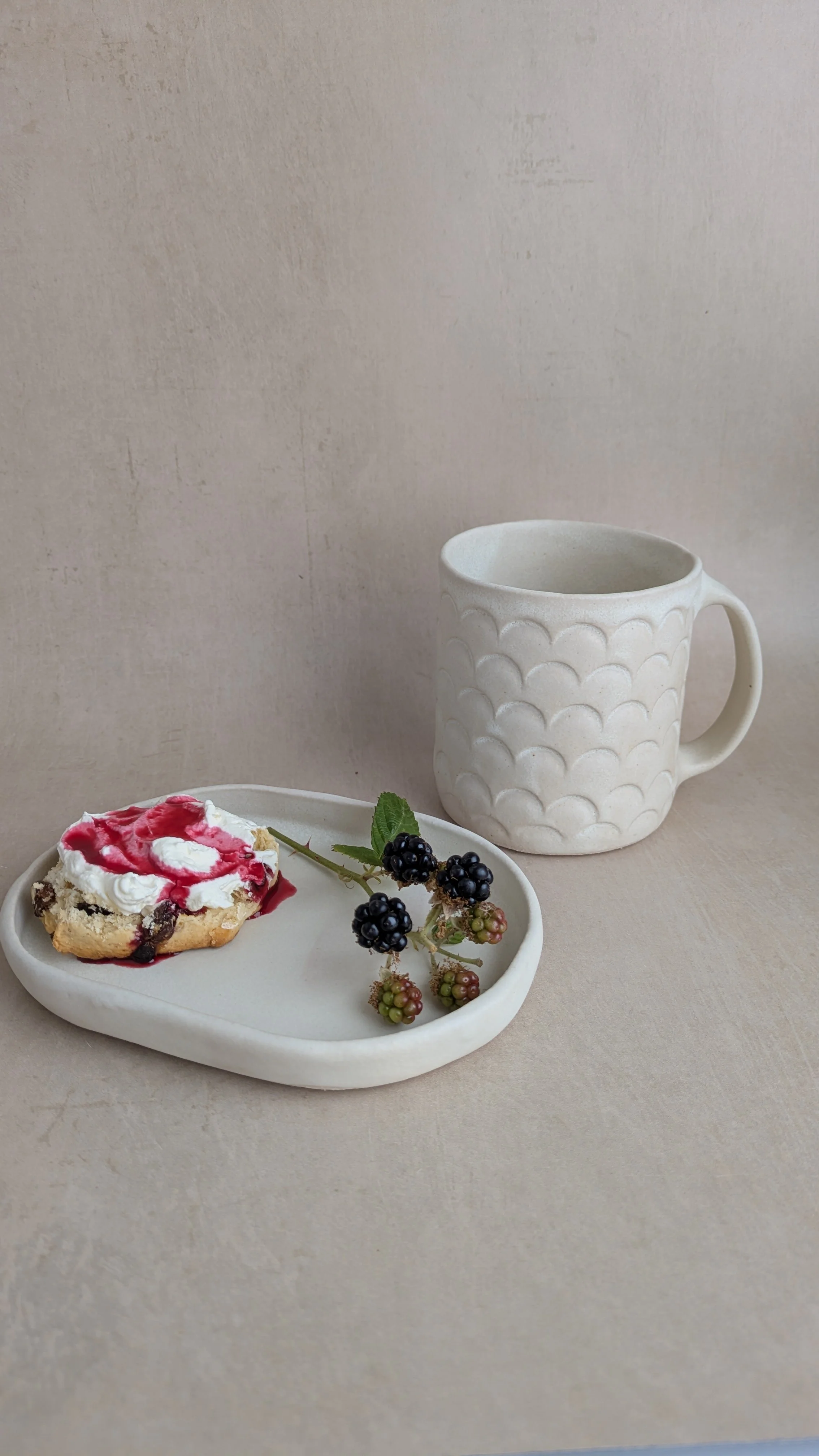 Ceramic scallop-pattern mug and plate with scone and blackberries arranged on a soft beige surface.