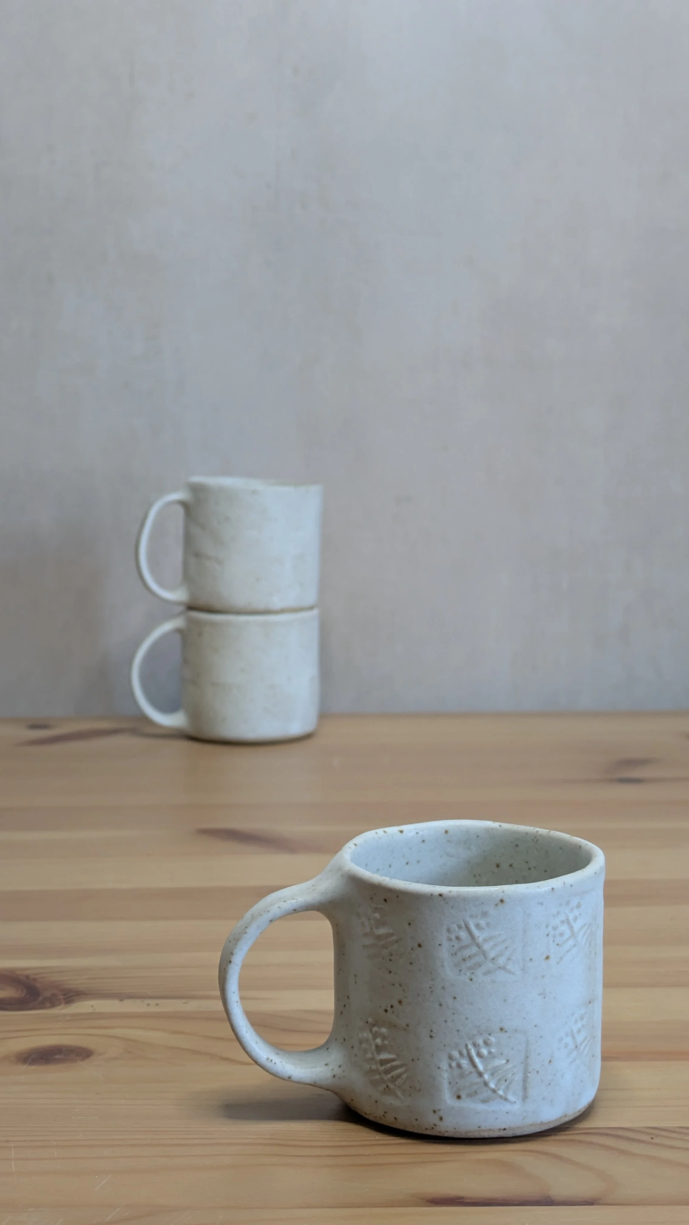 Handcrafted cream-glazed ceramic mug with subtle leaf impressions, displayed on a wooden table with two stacked mugs in the background.