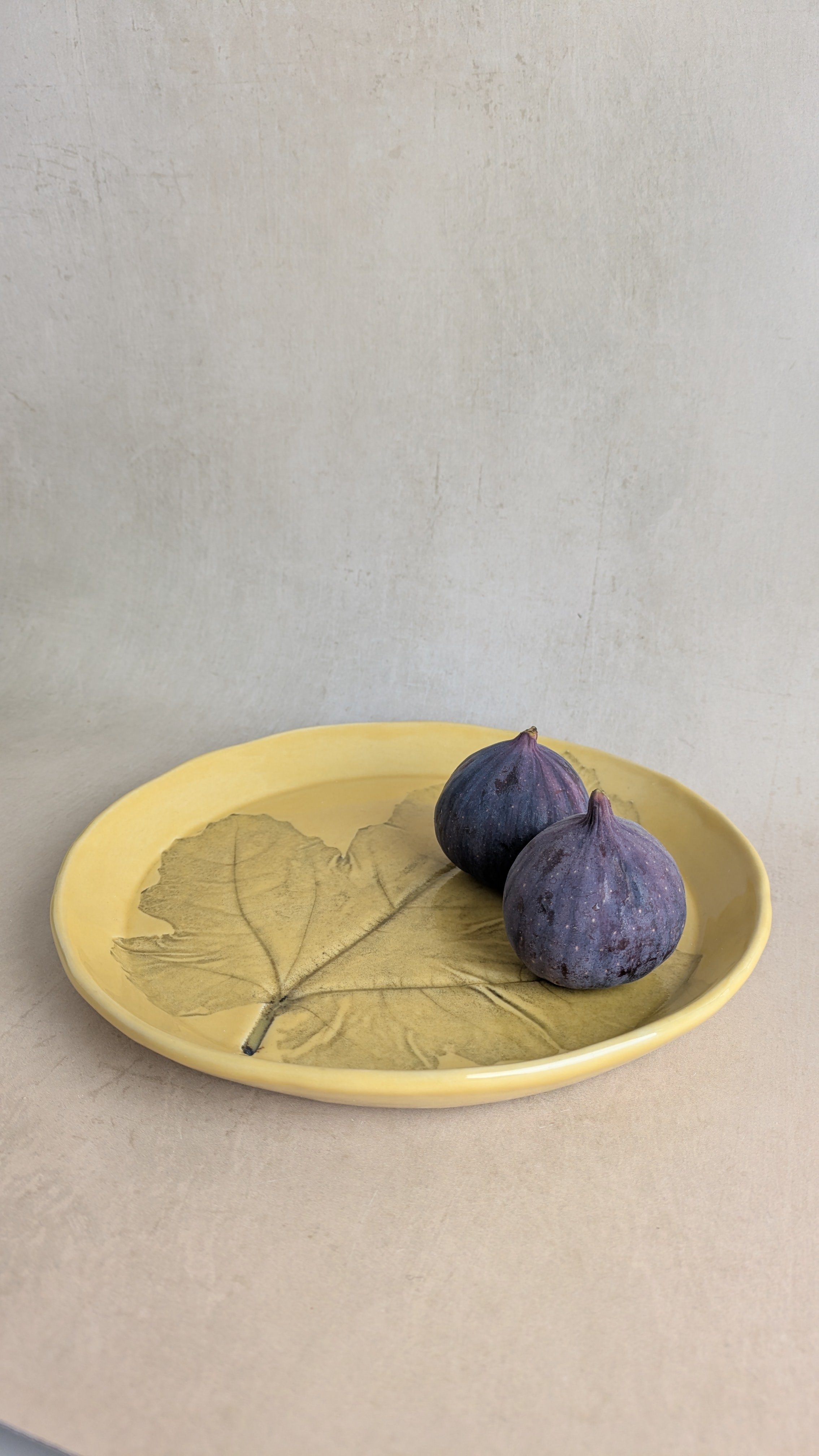 Handmade yellow‑glazed stoneware plate with fig‑leaf imprint, styled with mug and figs.