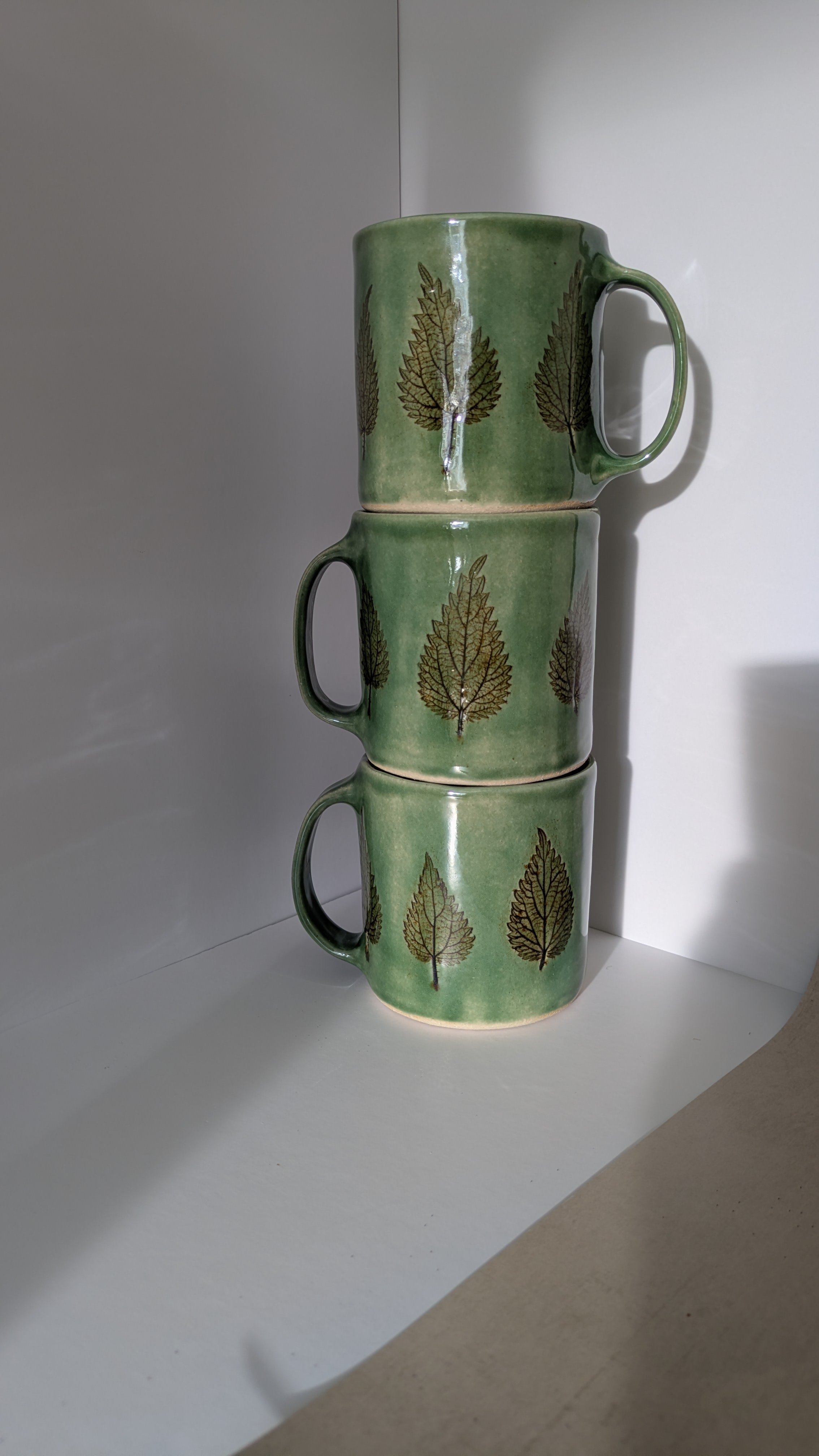 Three large green stoneware mugs stacked vertically, each with pressed nettle leaf motifs and glossy glaze.
