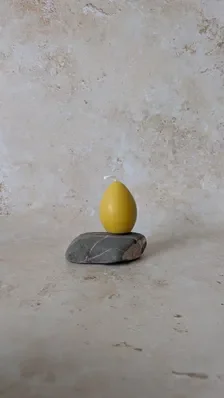 Hand‑poured yellow beeswax egg shaped candle resting on a smooth grey stone with white streaks, set against a soft beige textured background for a simple, natural composition.