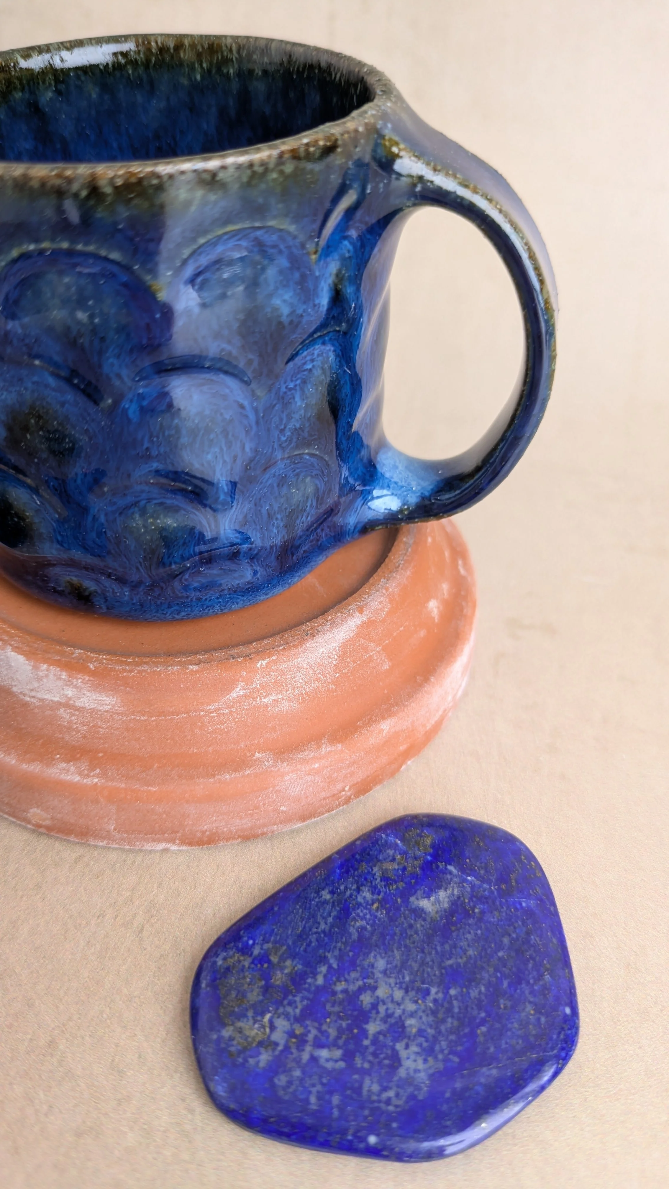 Close-up of a glossy blue arch-textured mug resting on a terracotta saucer with a lapis lazuli stone in front.