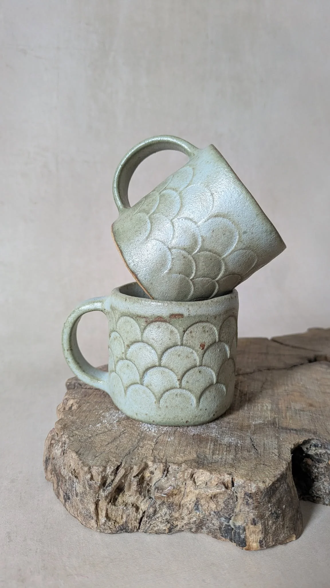 Two handmade sage‑green ceramic mugs with scalloped texture, stacked on a piece of natural wood, crafted at Fireweed Pottery.