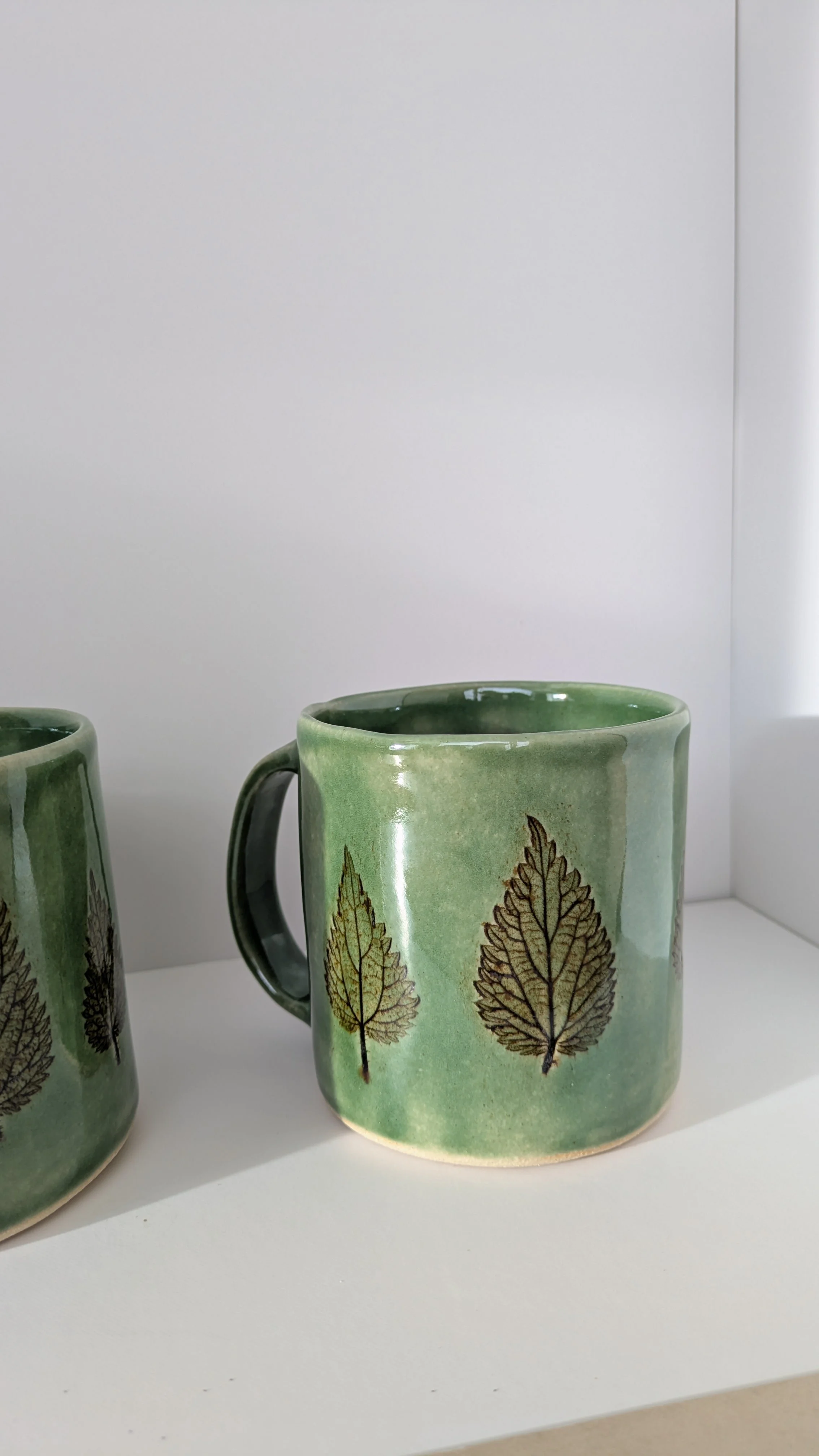 Two green stoneware mugs with nettle leaf design, side by side on a white background.