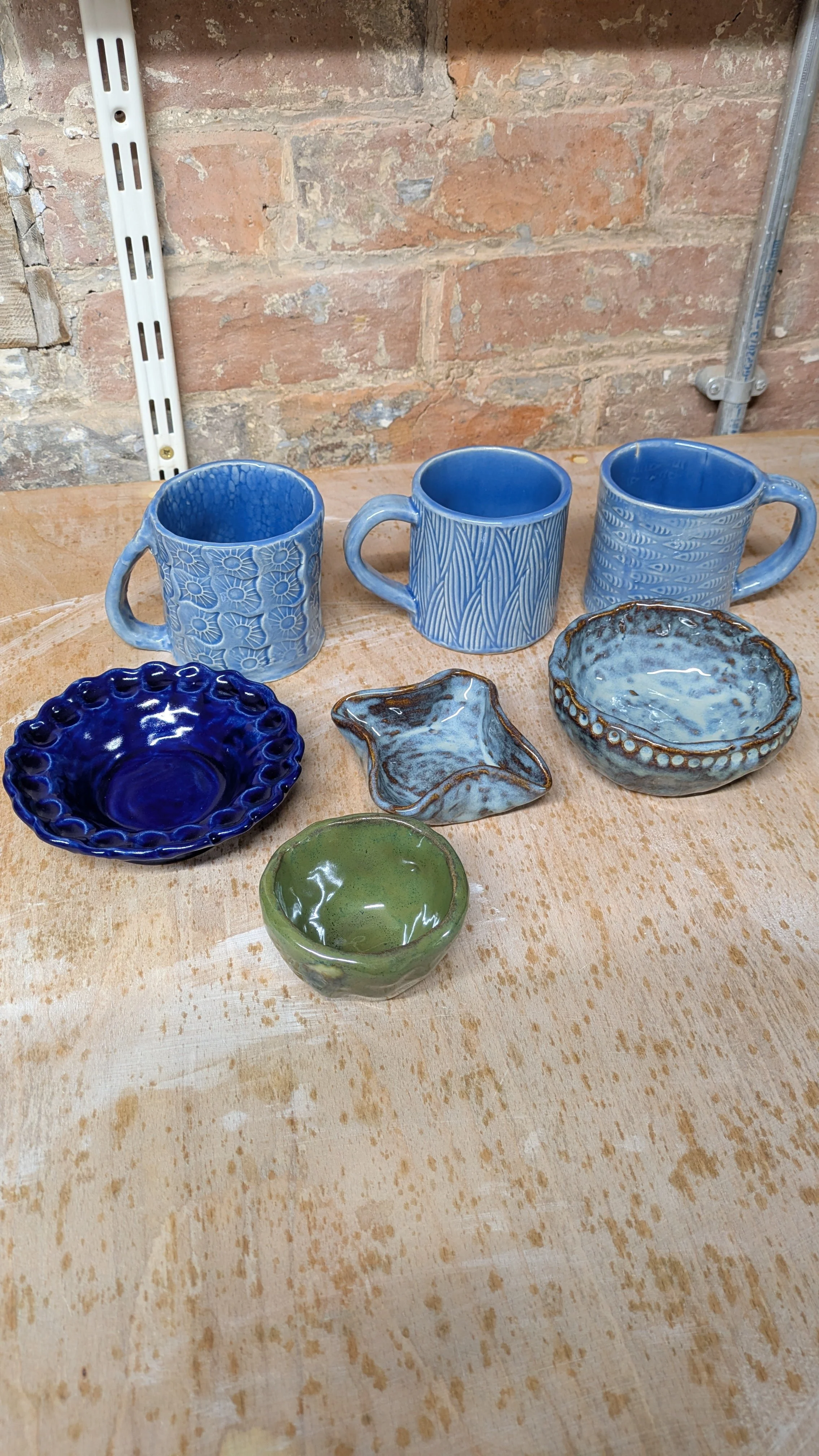 Collection of finished handmade pottery pieces including blue mugs, bowls, and dishes displayed on a wooden table against a brick wall.