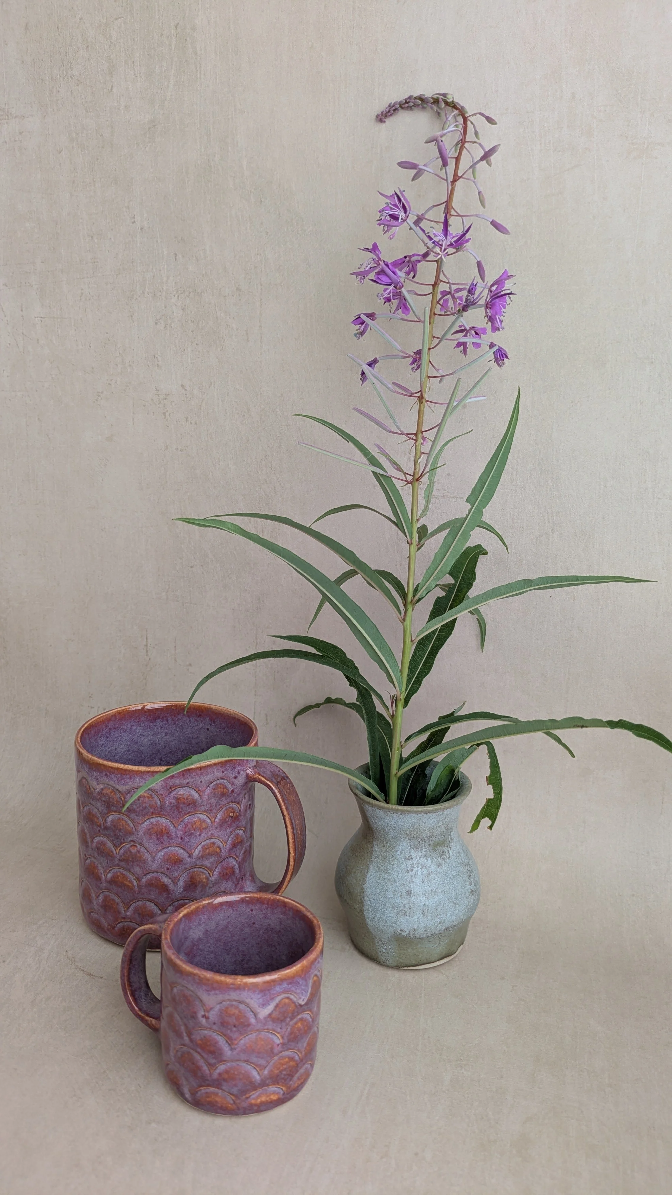 Two textured purple mugs and a small vase with a fireweed stem, displayed on a neutral background.