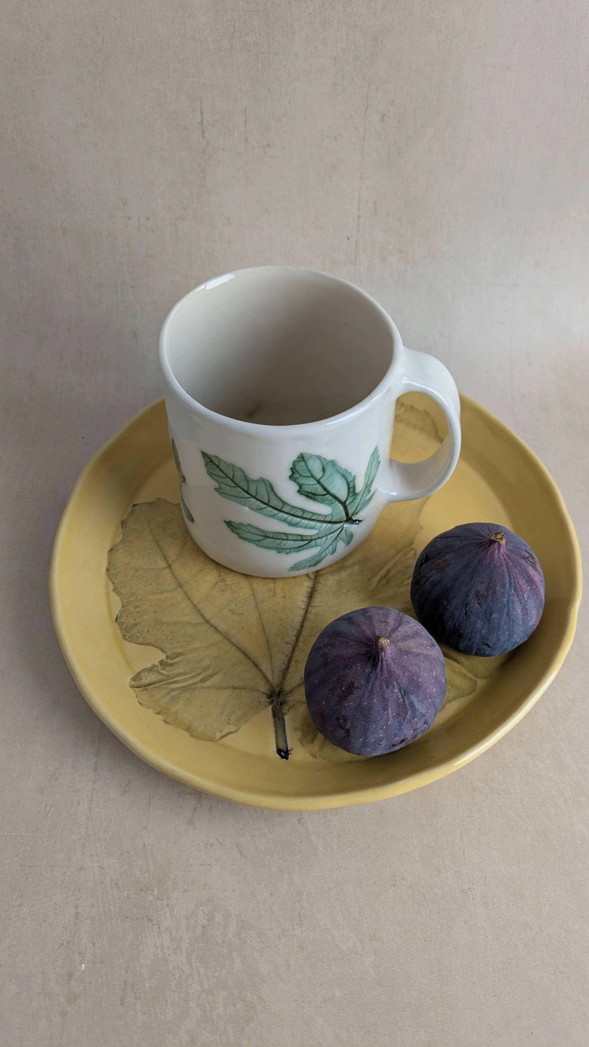 Ceramic plate with fig‑leaf design and white mug, styled with two purple figs on a neutral background.