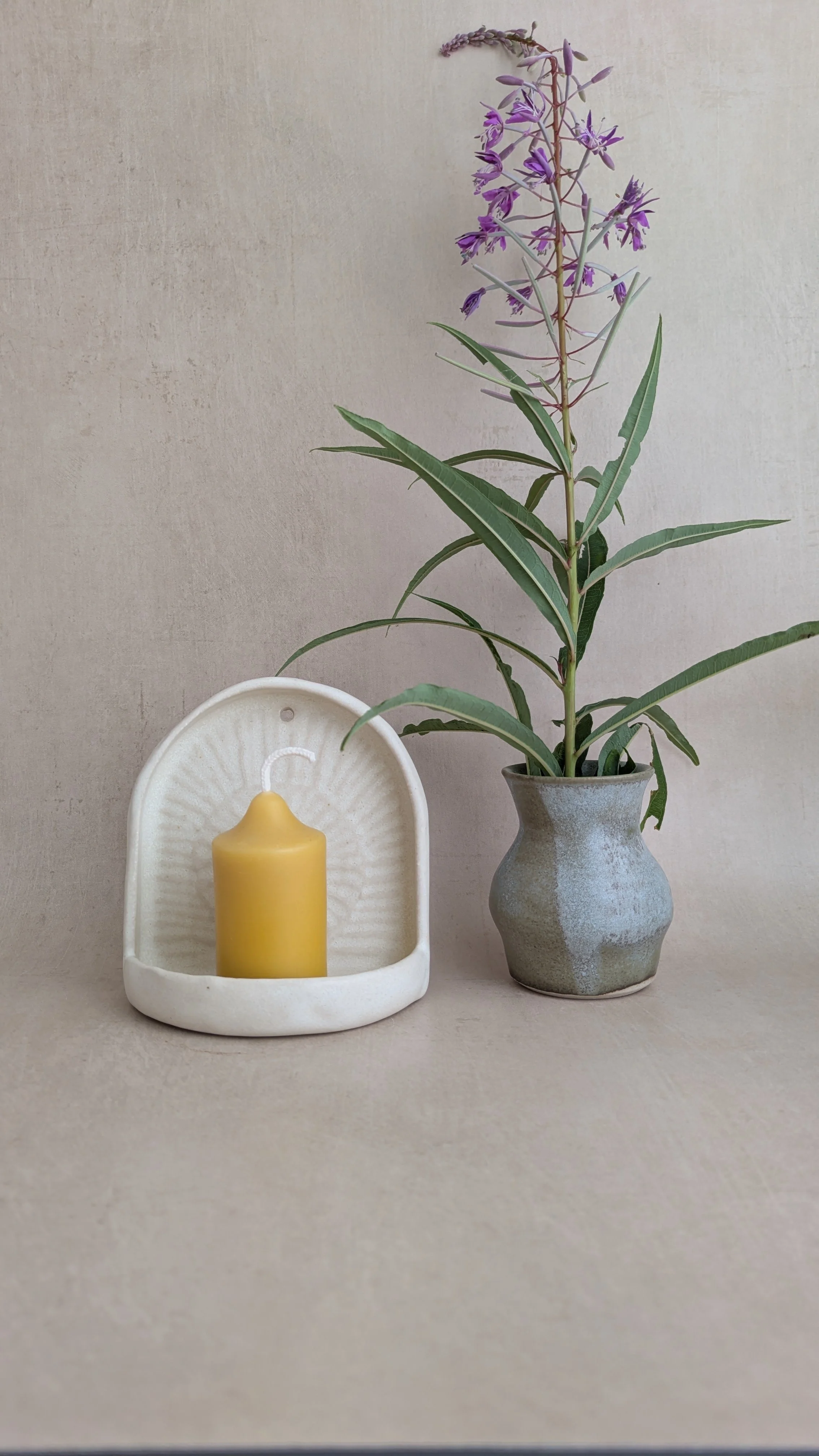 Cream ceramic wall sconce with beeswax candle beside a handmade vase holding fireweed flowers.