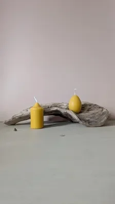 Pillar and egg beeswax candles displayed together on a curved piece of driftwood against a soft pink wall, styled simply and naturally.