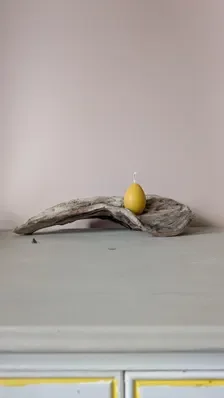 Single handmade yellow beeswax egg‑shaped candle resting on a curved piece of driftwood, styled on a pale grey surface with a soft pink wall behind, creating a calm, natural composition.