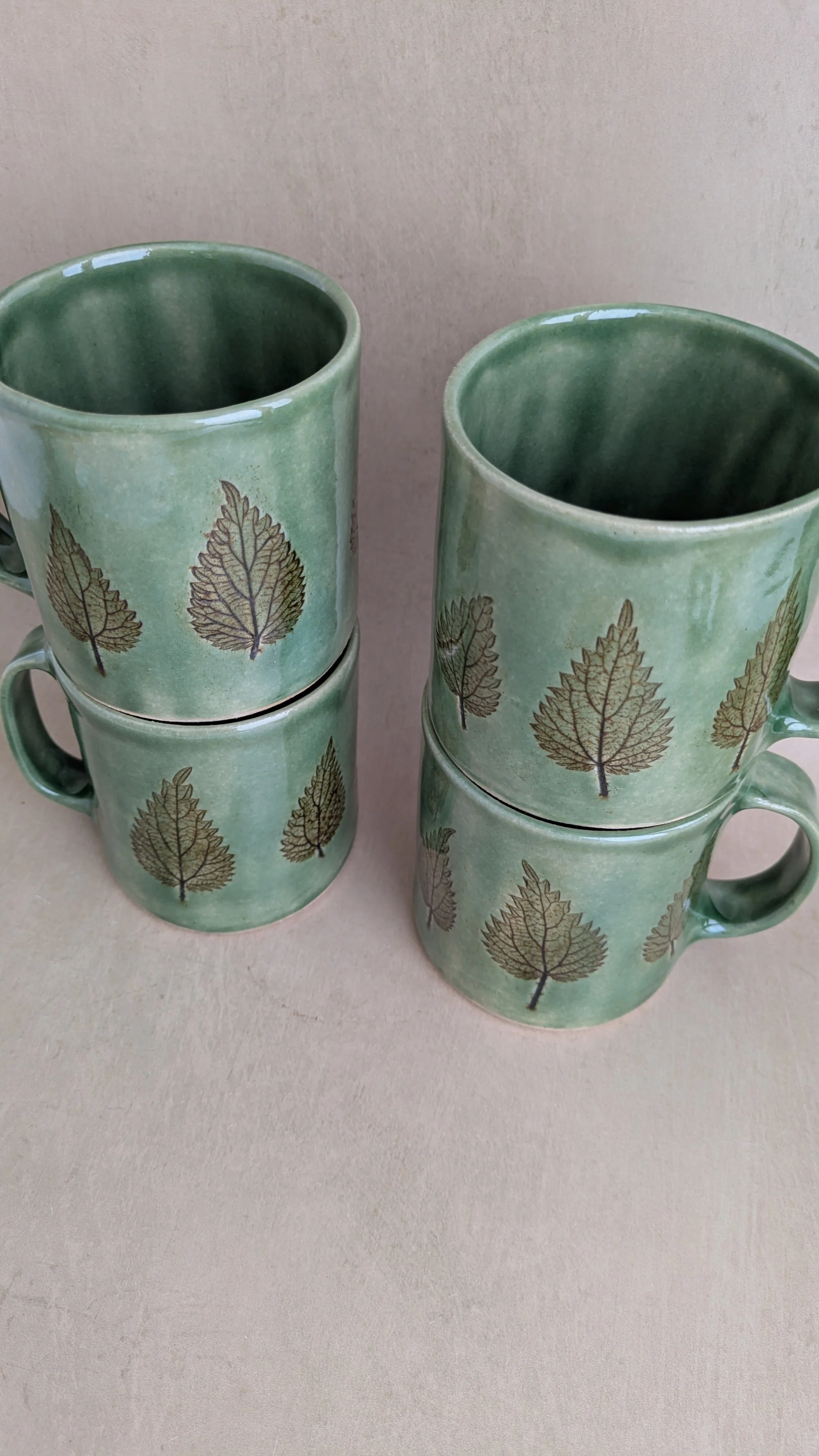 Four large nettle mugs arranged in two stacks on a neutral surface, showing botanical leaf pattern and green glaze.