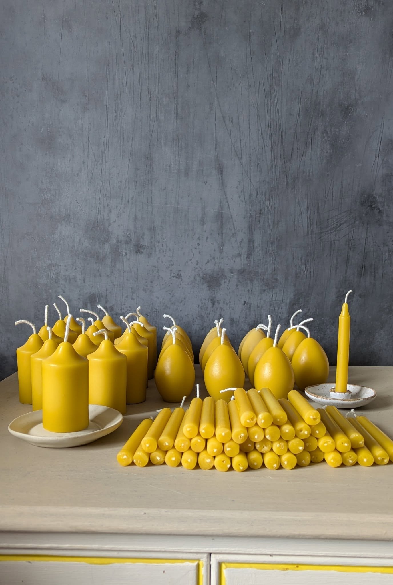 Display of handmade beeswax candles in various shapes and sizes arranged on a light surface against a textured grey background.