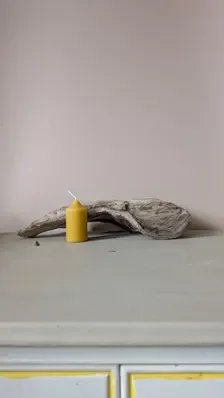 Pillar beeswax candle beside driftwood on a pale grey tabletop with a soft pink wall behind, highlighting its clean, simple shape.