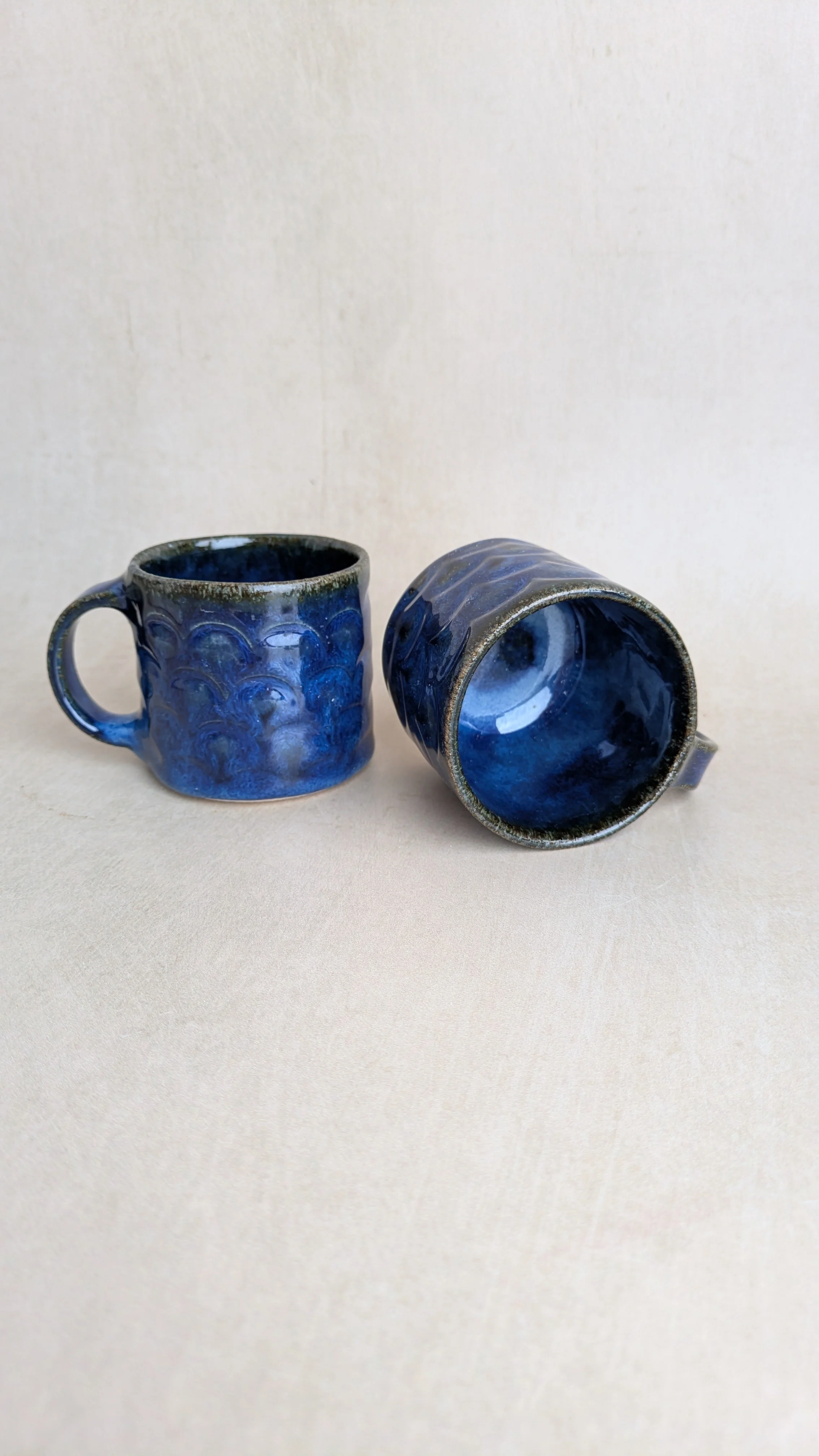Two Azul Lazuli mugs with scalloped texture, one upright and one lying on its side to show interior glaze.