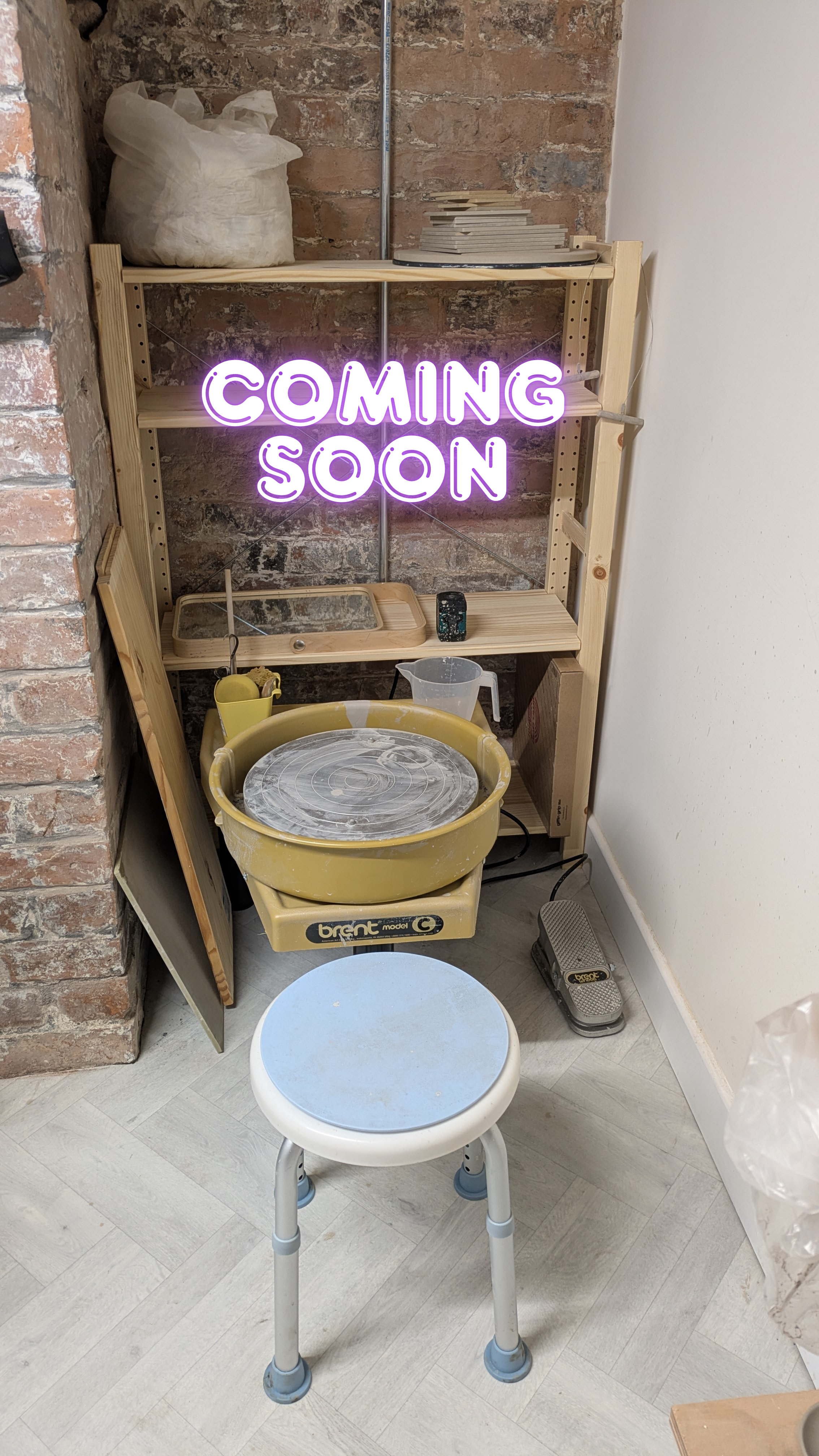 Compact pottery workspace featuring a yellow Brent Model C wheel, stool, and shelving with clay and tools against a brick wall.