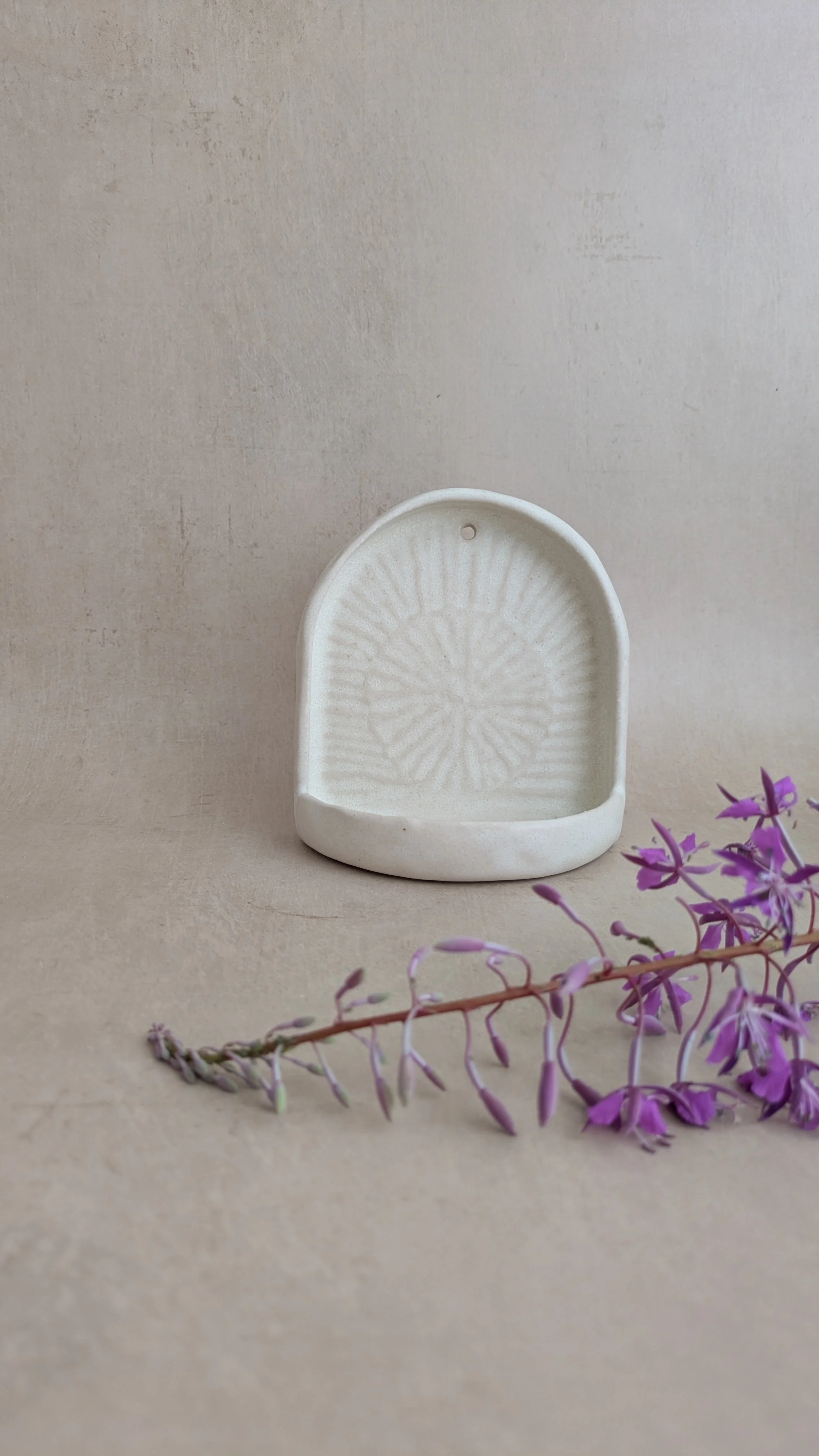 Cream arched ceramic wall sconce with sunburst texture, styled beside a fireweed flower stem.