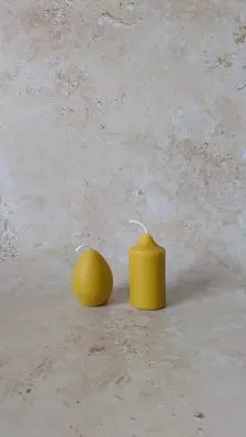Two hand‑poured yellow beeswax candles — one pillar and one egg‑shaped — displayed side by side on a light stone surface with a neutral beige backdrop, highlighting their warm tone and clean forms.