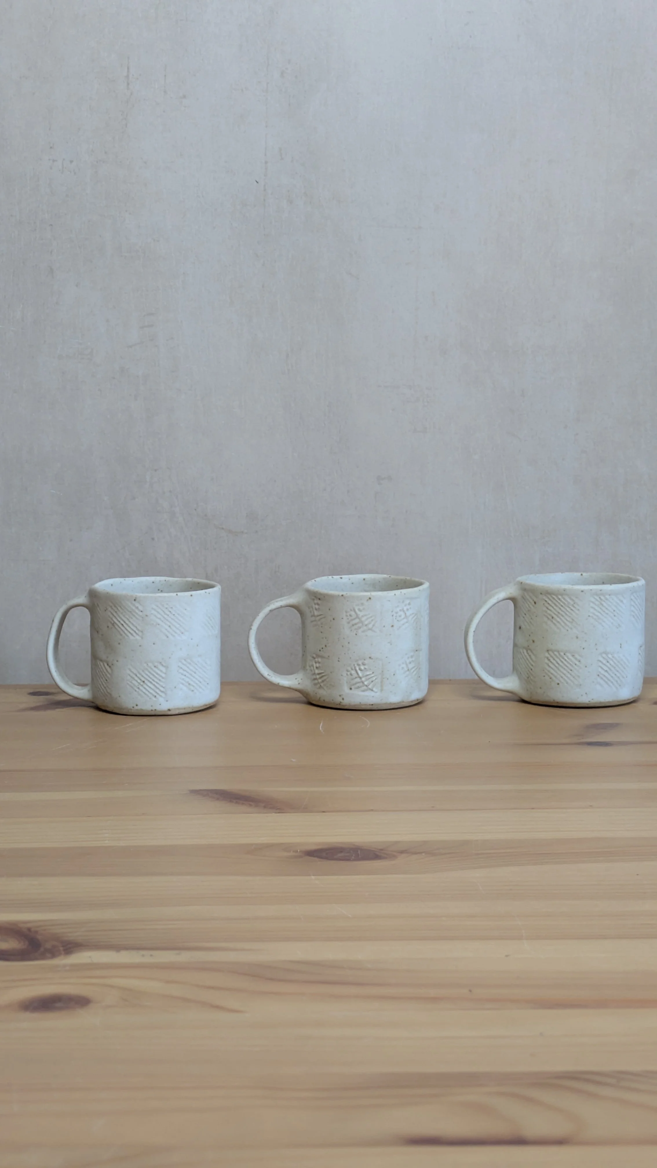 Three handcrafted cream-glazed ceramic mugs with subtle textured patterns, arranged in a row on a wooden table against a soft grey background.