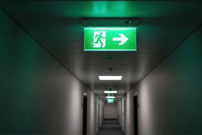 Green emergency exit sign with running figure and right arrow in a long, dimly lit corridor.